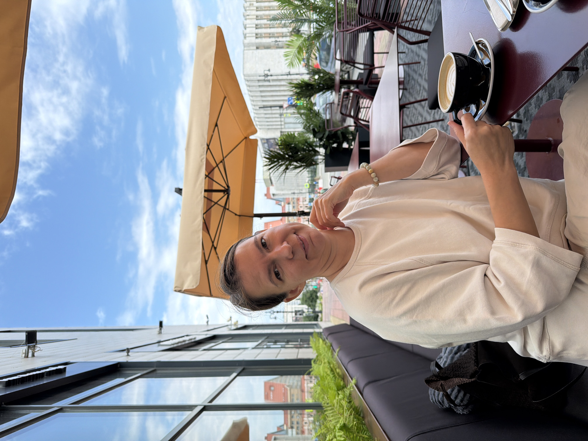 A woman sits at an outdoor table at a cafe in Perm, Russia. She is wearing a light-colored crew neck sweatshirt and pants. In front of her is a cup of coffee. The cafe has outdoor seating with tables and chairs, as well as umbrellas for shade. In the background, there is a building with large windows and some greenery. The sky is blue with some clouds.