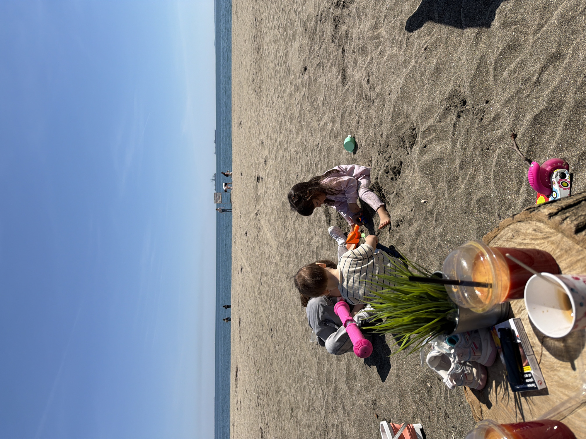 The image shows a beach scene in Unknown, Unknown, with two children playing in the sand. A girl in a pink shirt is bending over, while a child in a striped shirt is sitting and playing with beach toys. Nearby, there are drinks, a pack of cigarettes, shoes, and a small plant on what appears to be a tree stump. The beach has dark sand, and the ocean is visible in the background with a few people in the distance. The sky is clear and blue. A shadow is cast in the upper right corner of the image.