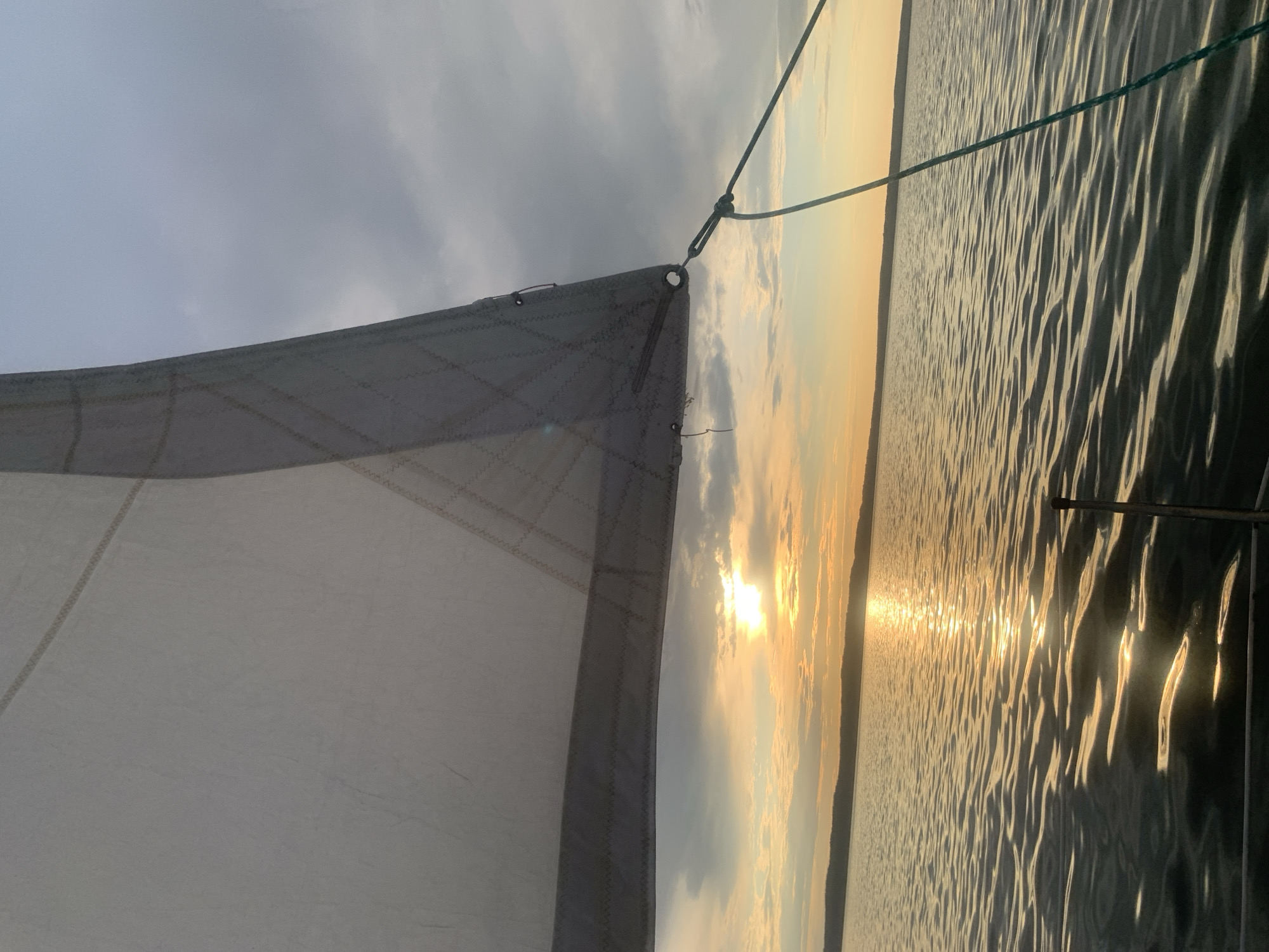 The image captures a serene moment on the water, likely a lake or a wide river, near Chaykovsky, Russia, as indicated by the presence of the Kama River. The perspective is from aboard a sailboat, with a large white sail dominating the upper left portion of the frame. The sail is partially obscuring a cloudy sky. The sun, low on the horizon, casts a warm, golden glow across the scene, its rays reflecting off the rippled surface of the water. The distant shoreline is visible as a dark, undulating line beneath the horizon. A green rope, attached to the sail, extends diagonally across the image, adding a nautical element to the composition. The overall atmosphere is tranquil and idyllic, evoking a sense of peace and natural beauty. There are no people visible in the image.