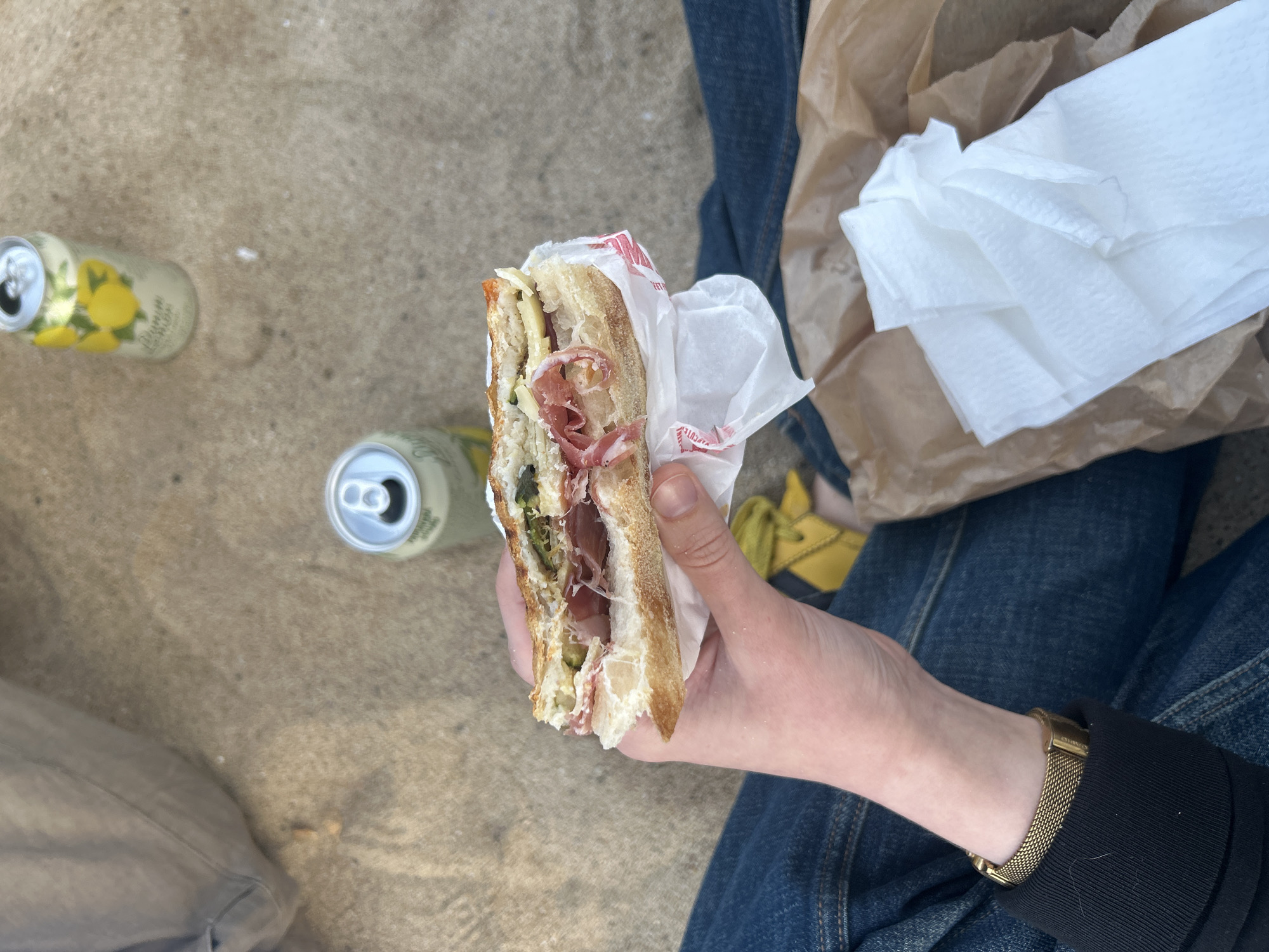 A person holds a half-eaten sandwich outdoors, with two cans of sparkling lemonade visible on the ground to the left. The sandwich is held in the left hand, partially wrapped in white paper with red print, and contains what appears to be meat, cheese, and greens. The person is wearing blue jeans and a black jacket, with a gold watch visible on their wrist. Next to them is a brown paper bag filled with white napkins. The background suggests an outdoor, urban setting, possibly in the city of Barcelona, Spain, based on the style of packaging and the informal, relaxed atmosphere. The ground is a light grey color, possibly concrete or stone.