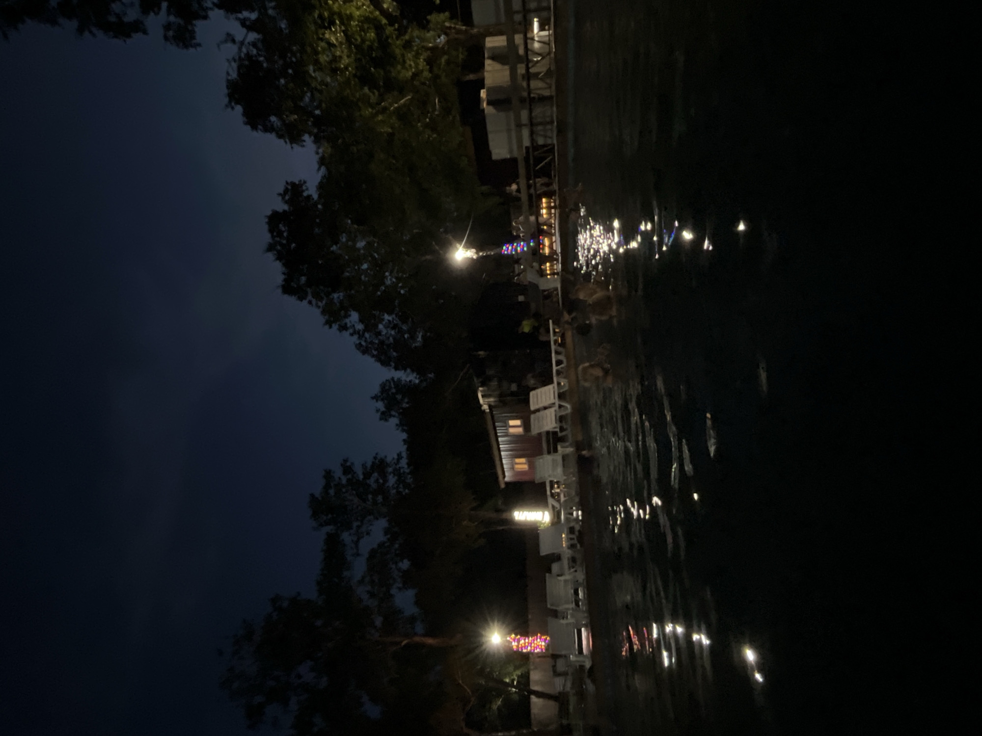 The image depicts a nighttime scene at a resort or hot spring facility in Paratunka, Russia. The foreground shows a dark body of water, likely a pool or thermal spring, with reflections of lights shimmering on its surface. Several lounge chairs are visible along the edge of the water, some illuminated by string lights that are multicolored. In the background, a wooden structure with windows appears to be a building, possibly a changing room or lodging. Trees surround the area, silhouetted against the dark night sky. A lamppost with a bright light is also present, along with a few people in the water and on the deck, though they are not clearly defined due to the low light conditions. The overall atmosphere is tranquil and suggests a recreational setting.