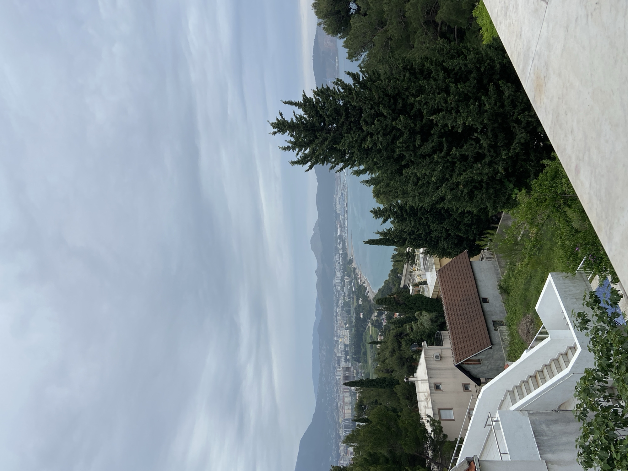 The scene is a view of Šušanj, Montenegro from an elevated perspective. The image includes the coastal city with its buildings, the Adriatic Sea, and the surrounding mountains under an overcast sky. In the foreground are residential buildings with tiled roofs and white stairs. Vegetation, including trees, is visible on the hillside, and architectural details such as balconies and walls are also visible. There are no people visible in the shot.