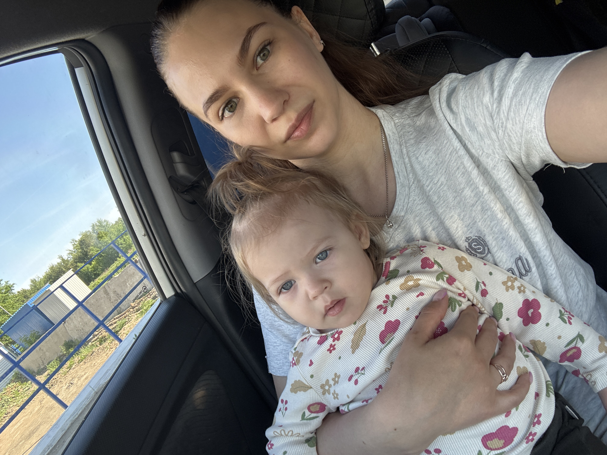 The image depicts a woman holding a baby, presumably in a car. The woman has her hair pulled back, light skin, and wears a grey t-shirt with a pendant. She is looking at the camera. The baby is held in her arms and is wearing a patterned, long-sleeved shirt. The baby has light skin and blue eyes. Outside the car window, there is a view of a blue sky, trees, a fence, and some buildings. Based on the visual clues of the buildings and fences, the location could possibly be Chaikovsky, Russia.