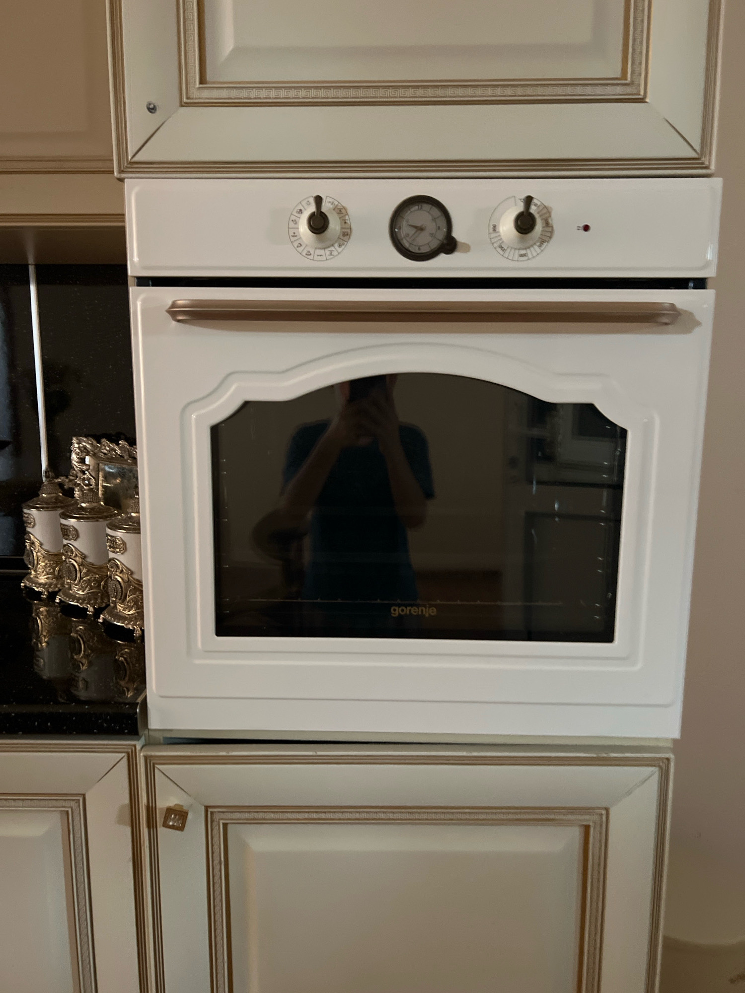 This is a medium shot, eye-level, indoor photograph of a white Gorenje oven. The oven is mounted above two cream-colored cabinet doors with gold trim. A dark countertop with three decorative canisters is visible to the left of the oven. The oven door has a glass window with the "Gorenje" logo. The oven's control panel features a clock and two knobs. A person is reflected in the oven door, wearing a blue shirt. The overall lighting is dim. The image is not specific to Novogagatli, Russia, or any particular location.