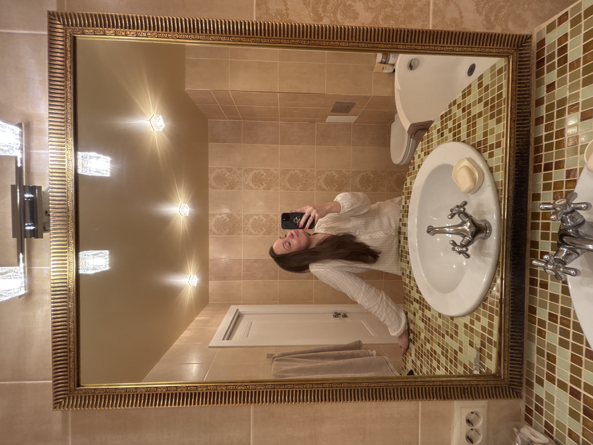 The image is a bathroom selfie taken in front of a large, ornate gold-framed mirror. The woman in the reflection is holding a smartphone and looking at its screen. The bathroom has beige tiled walls, a white door with a towel hanging behind it, and a sink with a soap dish on a mosaic countertop. A toilet is visible next to the sink. The ceiling has built-in lighting fixtures. It is likely a private bathroom in Chaikovsky, Russia based on the user’s location.