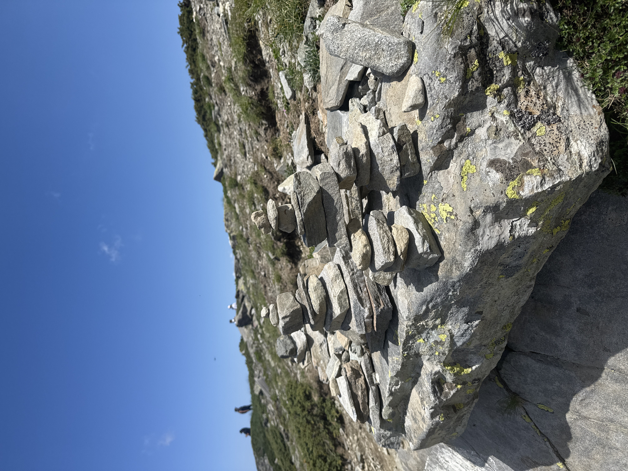 In Panichishte, Bulgaria, on a sunny day, a rocky mountainous landscape unfolds. In the foreground, several cairns, man-made stacks of stones, are prominently displayed on a large, rough-hewn boulder. The stones vary in size and color, with some exhibiting patches of yellow and green lichen. Behind the cairns, the terrain slopes upward, covered with sparse vegetation, rocks, and dirt. In the middle ground and background, a few individuals are visible, appearing as small figures walking on the hillside. The sky above is a clear, vibrant blue with a few wispy white clouds. The scene captures a serene mountain environment, possibly a hiking trail or a scenic overlook.