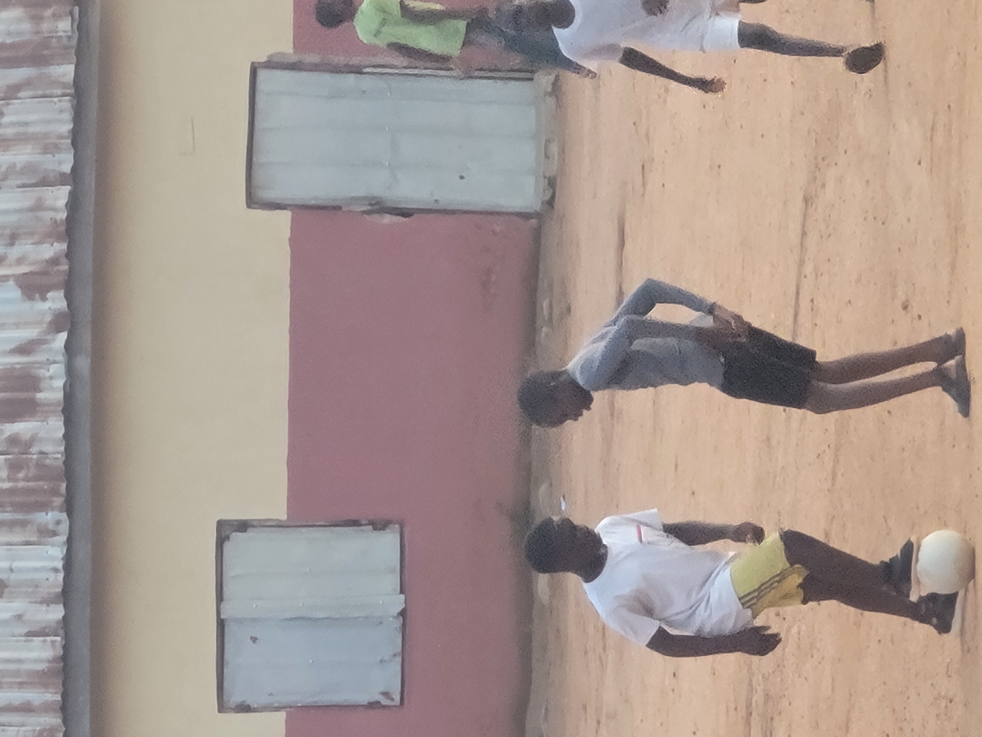The scene depicts an outdoor gathering in Barakin Akawo, Nigeria, where four dark-skinned individuals, appearing to be young males, are present on a reddish-brown dirt ground.
In the left foreground, one male, wearing a white short-sleeved t-shirt with a red-striped pocket and yellow shorts with dark stripes, controls a white soccer ball with his left foot. He wears dark shoes and faces towards the right. Adjacent to him, in the center foreground, a second male in a light gray long-sleeved shirt and dark shorts stands, facing partially left and looking downwards. He wears dark sandals.
In the right mid-ground, a third male, visible from the waist down, wears a white t-shirt and white shorts, and moves towards the left. He wears dark sandals. Further back on the right, a fourth individual is partially visible, wearing a bright green short-sleeved t-shirt and dark pants, walking towards the right.
The background consists of a two-toned building. The lower portion of the wall is reddish-brown, and the upper portion is light beige. A weathered, corrugated metal roof, showing signs of rust, covers the structure. On the left side of the building, a rectangular window opening is covered by two light-colored, possibly metal, panels within a dark frame. On the right side, a rectangular doorway contains a closed, light-colored door set within a dark frame. The ground is unpaved, consisting of loose dirt and small scattered debris.