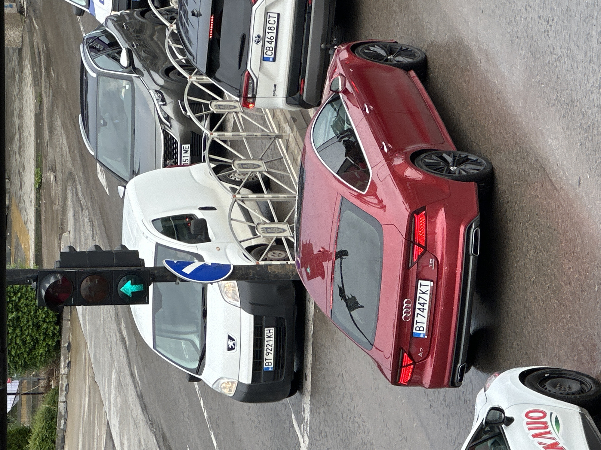 The image depicts a daytime street scene in Veliko Tarnovo, Bulgaria, under wet conditions, suggesting recent or ongoing rain. Multiple vehicles are stopped, likely at a traffic light, which displays a green arrow indicating a permitted left turn.
In the foreground, a red Audi A7, bearing the Bulgarian license plate "BT 7447 KT", is prominent. To its left, a white Peugeot Partner van with license plate "BT 9221 KH" is also stopped. Further back, a grey Toyota SUV is visible with the license plate "CB 46 18 CT". Another dark-colored vehicle, partially obscured, has the license plate "151 ME". On the right side, the front part of a white van with "KAUO" written on its side can be seen.
A series of metal bike racks are positioned in the median, separating lanes of traffic. The road surface is visibly damp, reflecting the overcast sky. A blue circular road sign with a white arrow, indicating a right turn or similar instruction, is also mounted near the traffic light. The scene captures typical urban traffic conditions on a wet day.