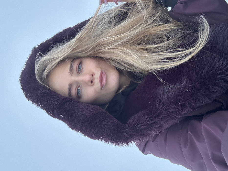 An outdoor close-up shot depicts a female subject from the chest up, positioned against an overcast or snow-reflected light gray sky in an unknown city and country. The subject, a fair-skinned individual with long, wavy blonde hair parted on the right side of her head, has blue eyes and a slight pink tint to her eyelids. Her lips are pursed forward. She is wearing a dark purple or maroon quilted winter coat featuring a prominent, fluffy dark purple faux fur-lined hood, which is pulled up and partially covers her hair. A black hooded garment is visible underneath the outer coat. Small white particles, consistent with snow or ice, are visible on the faux fur of the hood. The subject's left hand, with light-colored fingernails, is visible near her right shoulder, lightly touching or adjusting her hair. Her gaze is directed towards the camera.