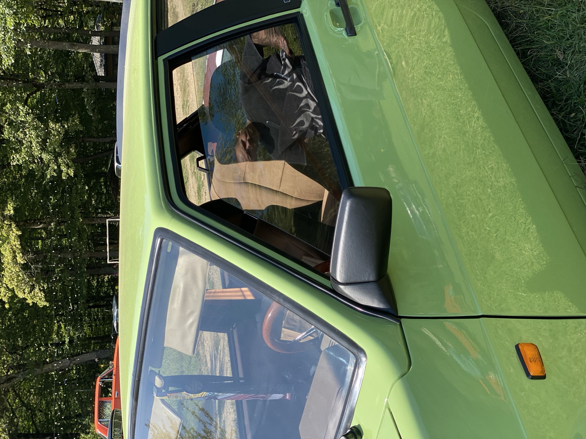 In Bezymyannoe, Russia, a bright lime green car, likely a Lada, is parked outdoors on a grassy area. Sunlight reflects off its polished exterior. The car's interior is partially visible through the open driver's side window. A tan-colored seat and a wooden steering wheel can be seen. A person wearing a black t-shirt with a flame design is reflected in the window, appearing to be seated inside. In the background, a line of trees forms a dense forest. An orange car is parked behind the lime green car. The overall scene suggests a casual outdoor gathering or event.