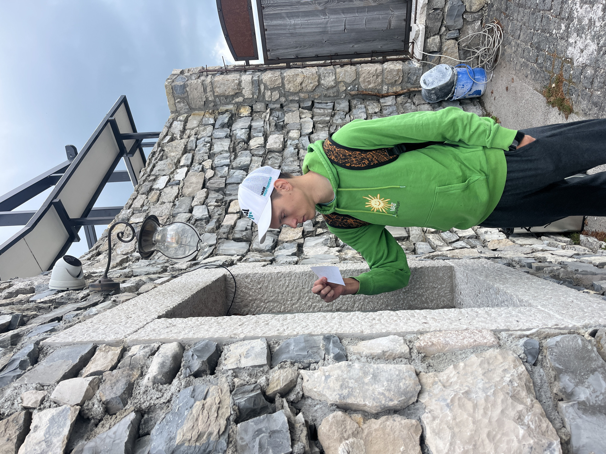 A young man in a green hoodie and white cap stands in Dolovi, Montenegro, holding a white card over a stone structure with a rectangular opening. The structure is built into a stone wall. A vintage-style light fixture is affixed to the wall near the opening. A security camera is also mounted on the wall. Gray skies can be seen in the background.