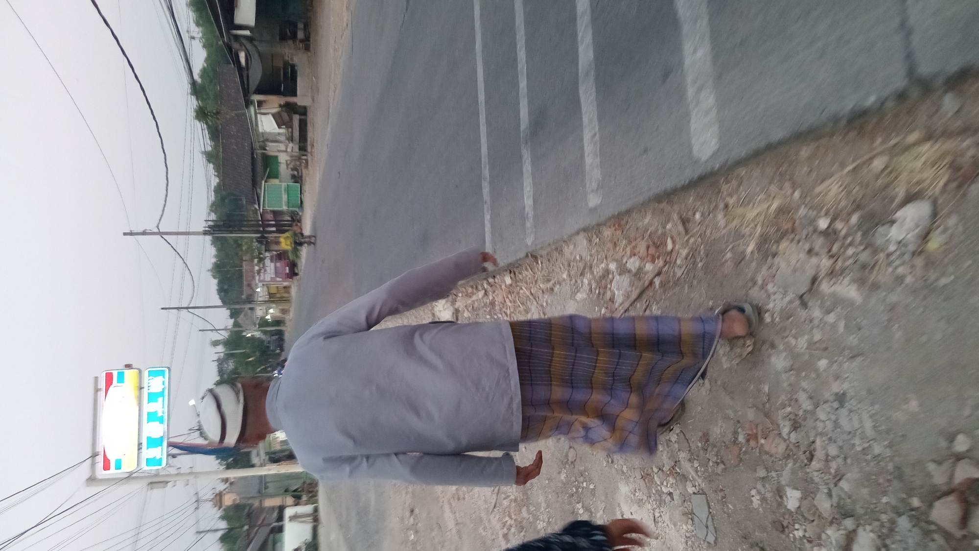In Trembulrejo, Indonesia, a man wearing a white peci hat, a light gray long-sleeved shirt, and a sarong with horizontal stripes of purple, yellow, and white walks along a dirt path next to a paved road. He is walking away from the camera, with his right arm slightly extended. To his left, another person's arm is partially visible. On the road, a pedestrian crossing with white stripes is marked. Further down the road, a person on a bicycle is visible. The background shows a collection of buildings and trees under an overcast sky. Electrical wires are strung overhead.