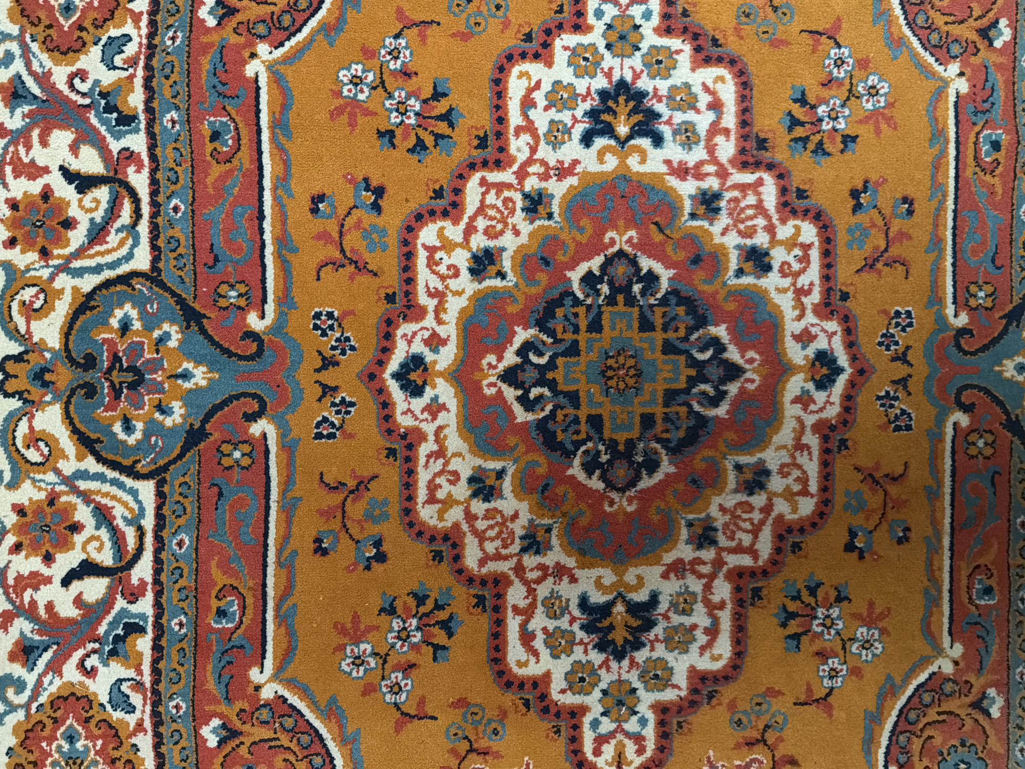 This close-up image presents a detailed view of a colorful, patterned rug or carpet. The primary subject is the textile itself, showcasing its intricate design and vibrant color palette.
The rug's central field features a warm, mustard yellow or golden background. Overlaid on this background are elaborate, traditional patterns, likely inspired by Persian or Oriental rug designs. A significant portion of a large, multi-layered central medallion is visible, characterized by complex floral motifs, stylized leaves, and geometric shapes. This medallion is rendered in a rich combination of colors, including off-white or cream, deep red or burnt orange, and various shades of blue, from light sky blue to dark navy or indigo. Black accents are used for outlining and adding depth to the details.
Surrounding the central field, distinct borders are partially visible on the left and right sides of the image. These borders also feature complementary patterns, including swirling arabesques, paisley-like elements, and small floral arrangements. Scattered across the mustard yellow field are delicate clusters of small flowers, often depicted with white and blue petals on slender stems.
The texture of the rug appears soft and woven, consistent with a textile with a visible pile. The lighting is even, suggesting an indoor setting. There are no people present, no specific activity taking place, and no visible text within the image. The rug appears to be in good condition. The location context provided is Засечное, Russia.