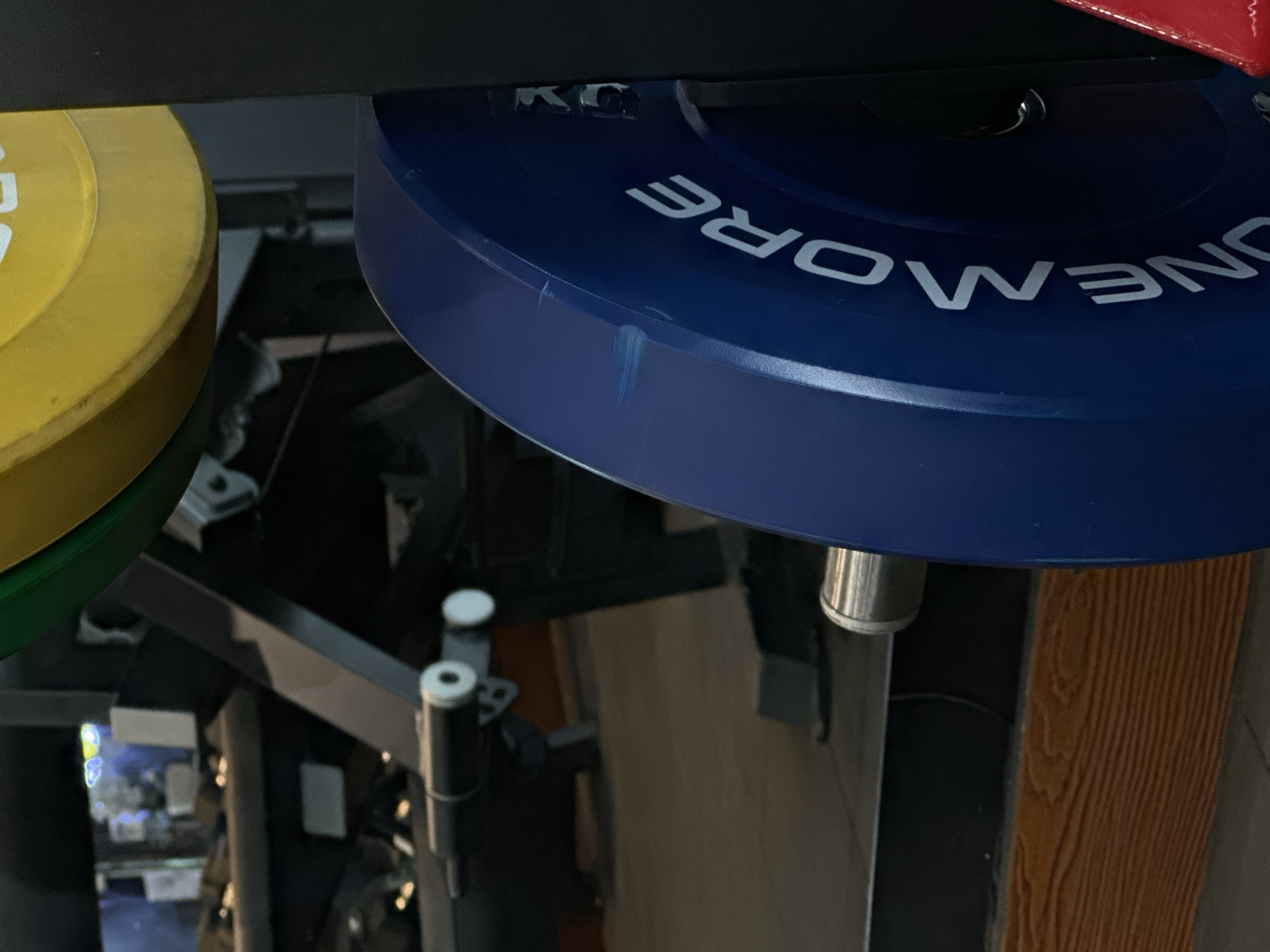 This close-up image, taken from a low angle, depicts multiple weight plates stored on a gym rack within an indoor fitness facility, potentially located in North Kuta, Indonesia. The most prominent object is a large, dark blue bumper plate in the foreground, featuring the brand name "ONEMORE" in white capital letters across its surface. Above this text, a small fragment of other white text, possibly indicating weight in "KG", is visible. To the left, a vibrant yellow weight plate is partially visible, and beneath it, a sliver of a green plate can be seen.
The plates are mounted on a dark metal rack structure. A silver, cylindrical sleeve for holding the plates is visible directly below the blue plate. In the lower-left, two vertical black metal posts with white caps are part of the rack's support system. A small red object is partially visible at the very top right. The background is somewhat blurred and dark, showing indistinct structural elements and equipment typical of a gym environment. A notable detail in the lower-right background is a vertical surface with a distinct brown wooden texture. No people or specific activities are depicted; the image focuses solely on the static gym equipment. The lighting appears to be artificial, typical of an indoor setting, with a faint light source visible in the far-left background.