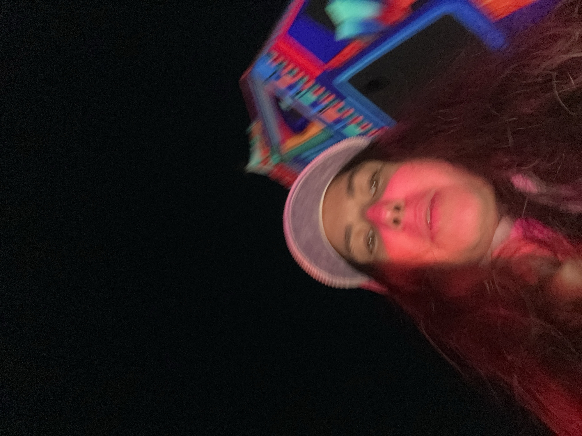 A low-angle, blurry photograph depicts a person with red hair and pink makeup on their face, wearing a pale-colored hat with a ridged brim. Above them, out of focus, is a multi-colored, angular structure with red, blue, turquoise, and possibly other colored geometric shapes. The background is primarily black, indicating it is likely nighttime. The photograph appears to be taken from a tilted perspective, looking upward. There are no other identifiable people, objects, or actions evident in the image. The city of Kazan, Russia, cannot be confirmed solely based on the visual information provided.