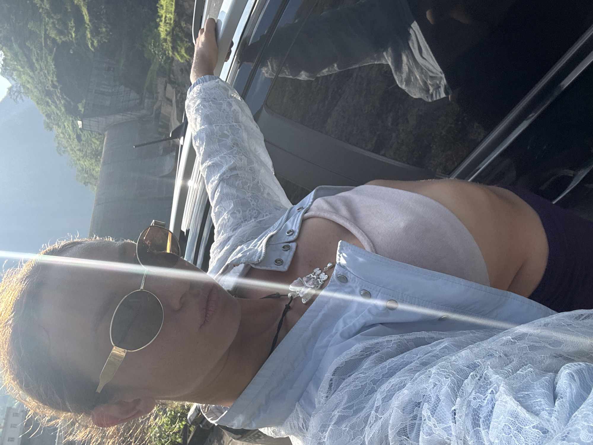 A woman in sunglasses and a blue lace top leans against a car. The background shows a green, hilly landscape, likely near a structure of concrete, possibly a dam, within the general vicinity of Sơn Vĩ Commune, Vietnam. The woman's hand rests on the top edge of the car door.