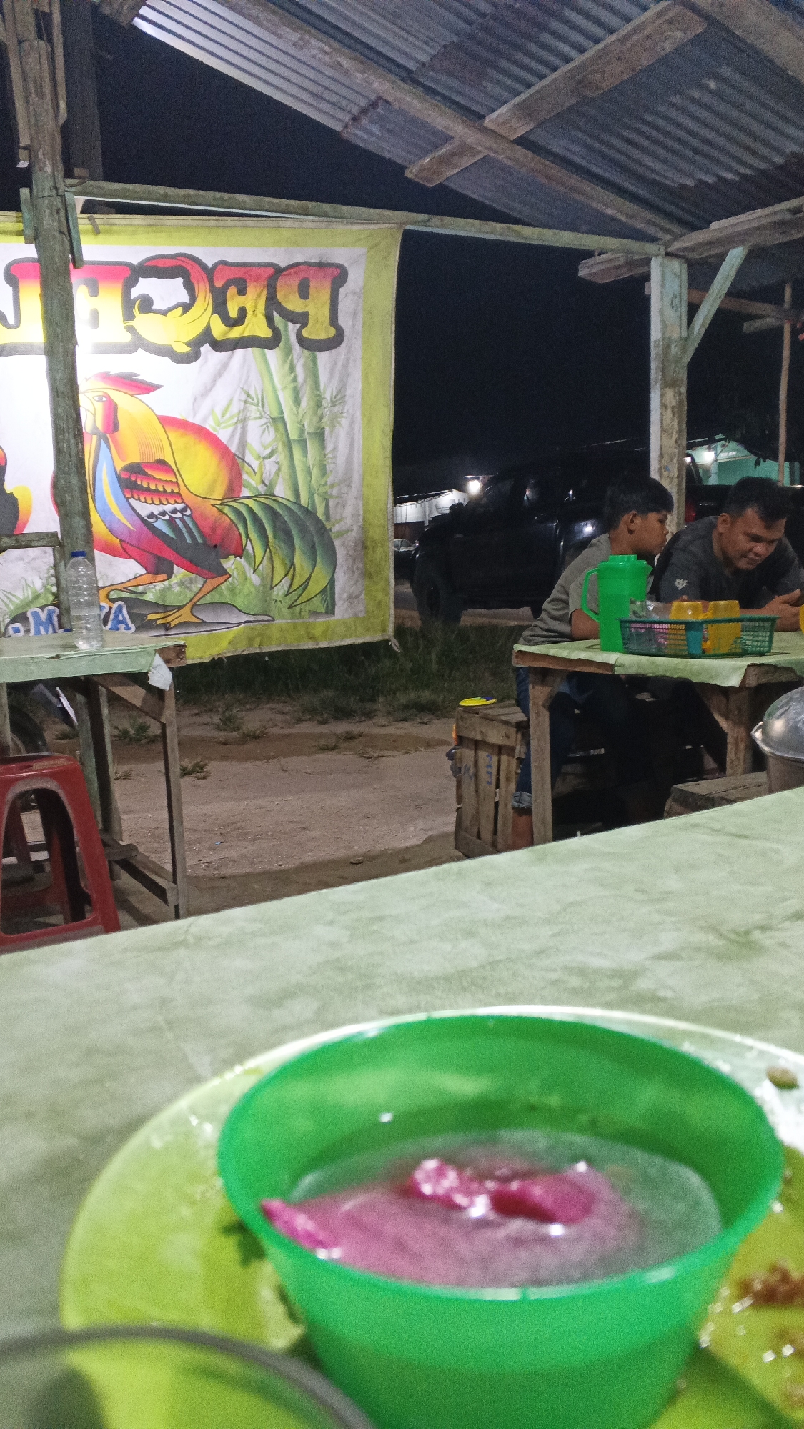 The image depicts a nighttime scene at an outdoor food stall, likely located in Tumang, Indonesia. In the foreground, a bright green plastic cup filled with a pinkish liquid and some solid chunks is visible on a green surface. This suggests a beverage or dessert is being served.
The mid-ground shows two individuals seated at a wooden table. One is a young boy wearing a grey t-shirt, looking towards the right. Beside him, an adult male, possibly his father, wearing a dark t-shirt, is also seated at the table, looking down and engaged with something on the table. A green basket containing yellow cups and other items sits on the table. Behind them, a dark-colored vehicle is parked.
The background features a large banner with a colorful depiction of a rooster and bamboo stalks, along with text that appears to read "PECA" in large yellow lettering with a red outline, and "M A" in smaller text. This banner likely advertises the name of the food stall or a specific dish. The stall is covered by a corrugated metal roof supported by wooden beams, creating a rustic ambiance. The ground appears to be dirt or gravel. The overall atmosphere suggests a casual, local eatery operating at night.