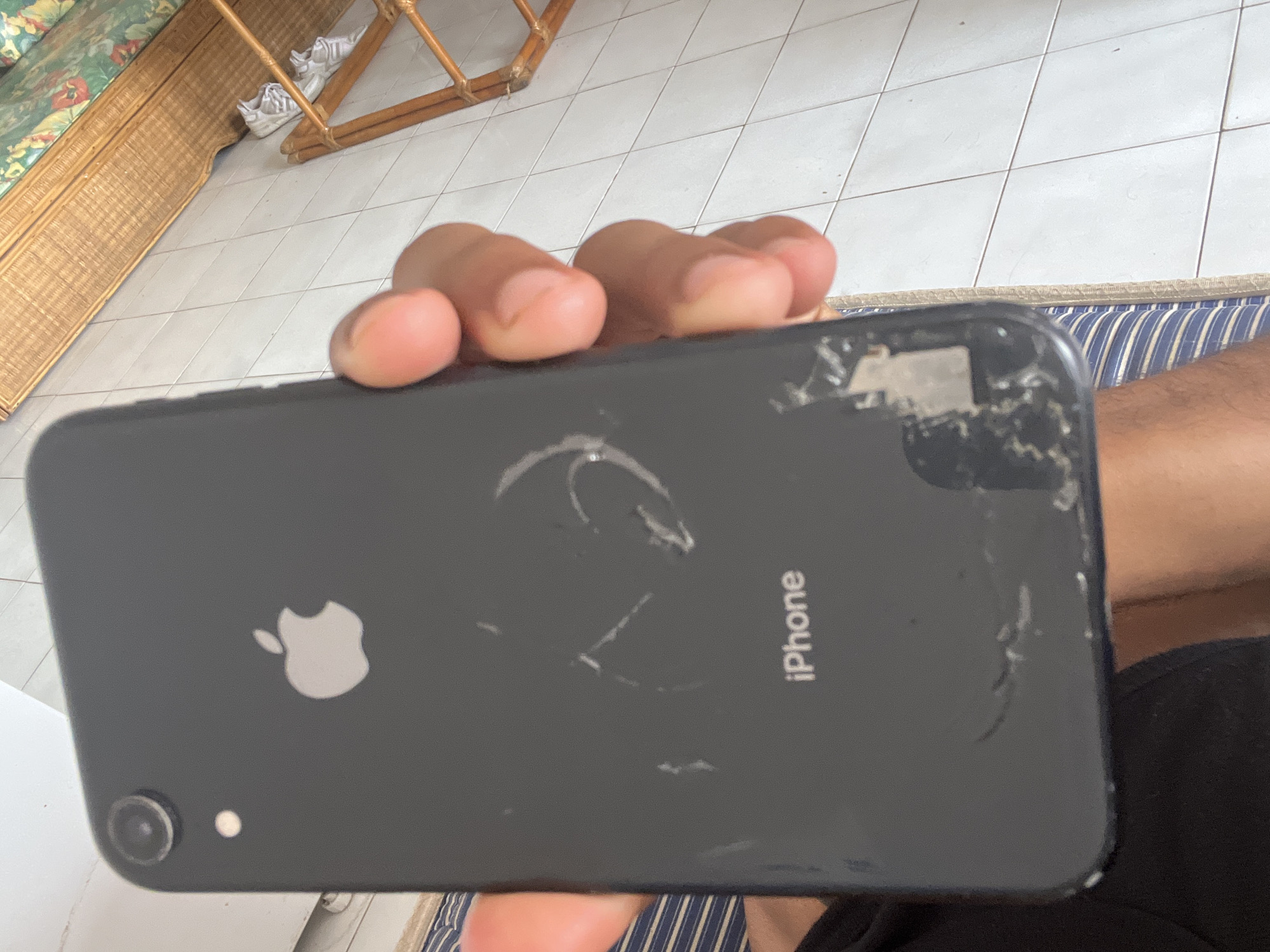 A person is holding a black iPhone in their right hand. The phone has significant damage to the back glass, particularly near the top edge and in a circular pattern around the Apple logo. The phone is being held indoors, and the background includes tiled flooring, a woven bench, and part of a blue and white striped cushion or surface. The location is likely Porlamar, Venezuela, but there are no elements in the image to confirm that.