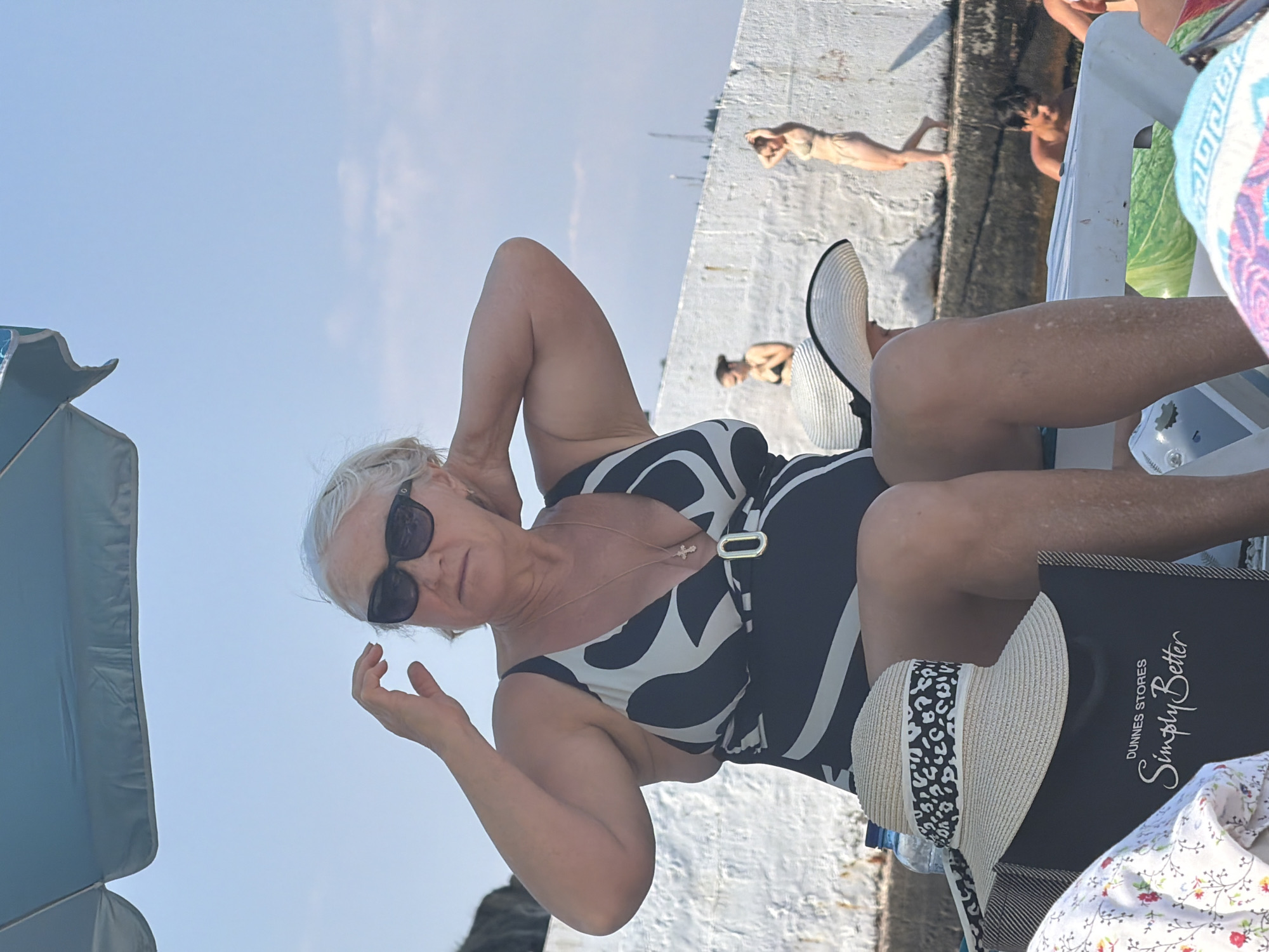 A middle-aged woman with gray hair and sunglasses is sitting on a beach chair under a blue umbrella, wearing a black and white patterned swimsuit. She is touching her hair with her hands. In the background, there are two other women in swimsuits standing near a white wall, and a young man is visible in a body of water. There are also sun hats and a beach bag in the foreground. The scene appears to be a sunny day at a beach or pool area, possibly in Partenit, Ukraine, given the coastal elements and architecture.