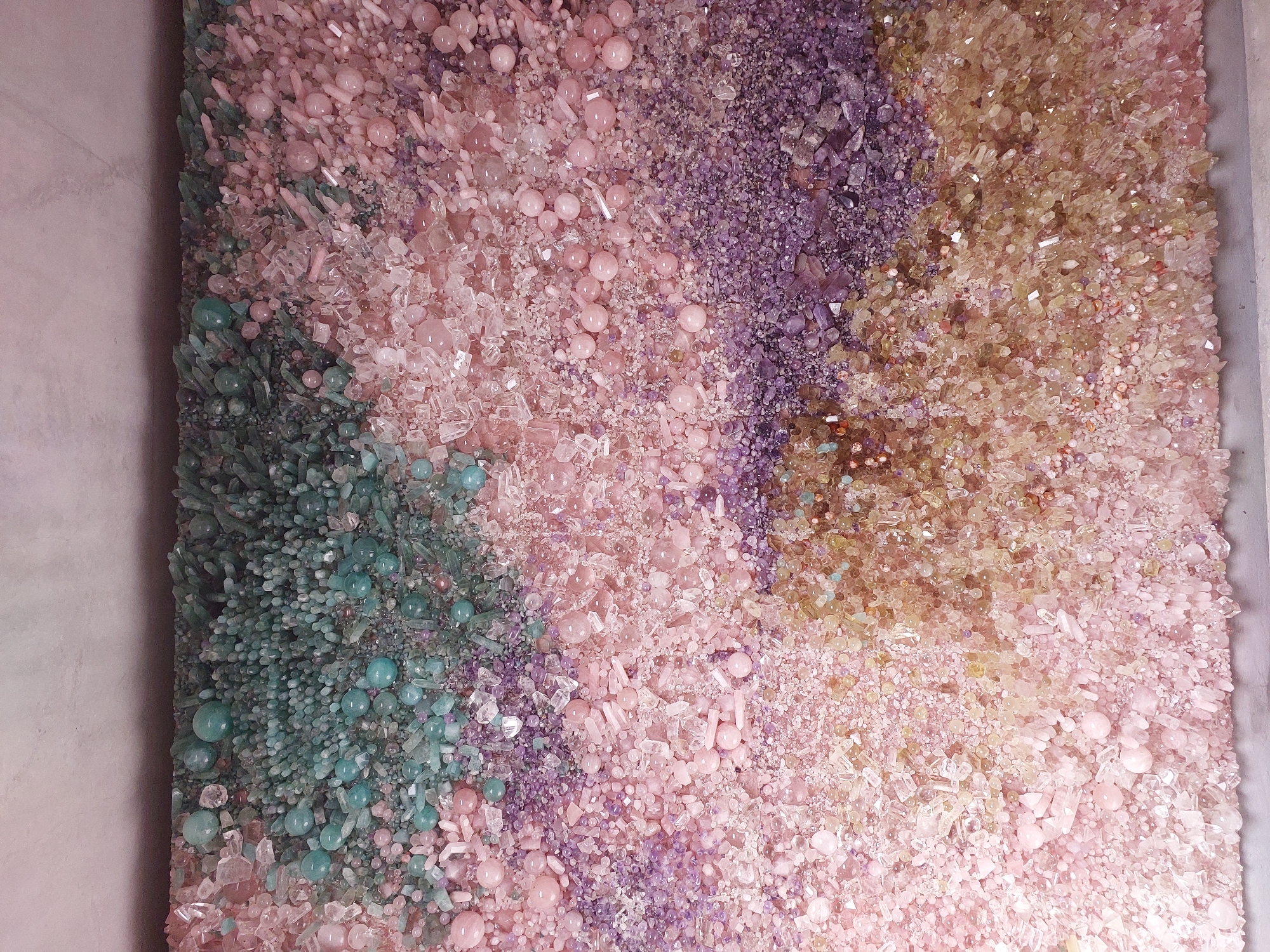 The image shows a close-up, overhead view of a large, textured artwork composed of numerous small, crystalline objects and smooth spheres in varying shades of pink, purple, teal, and pale yellow. The elements are densely packed, creating a mosaic-like effect with a sense of depth due to the uneven surfaces and varied sizes of the components. The artwork appears to be installed in a dimly lit, modern interior space, with a plain, light-colored wall or ceiling visible at the top. No people, specific actions, or interactions are depicted. The location in the city of Monaco is not discernable from the visual information provided in the image.