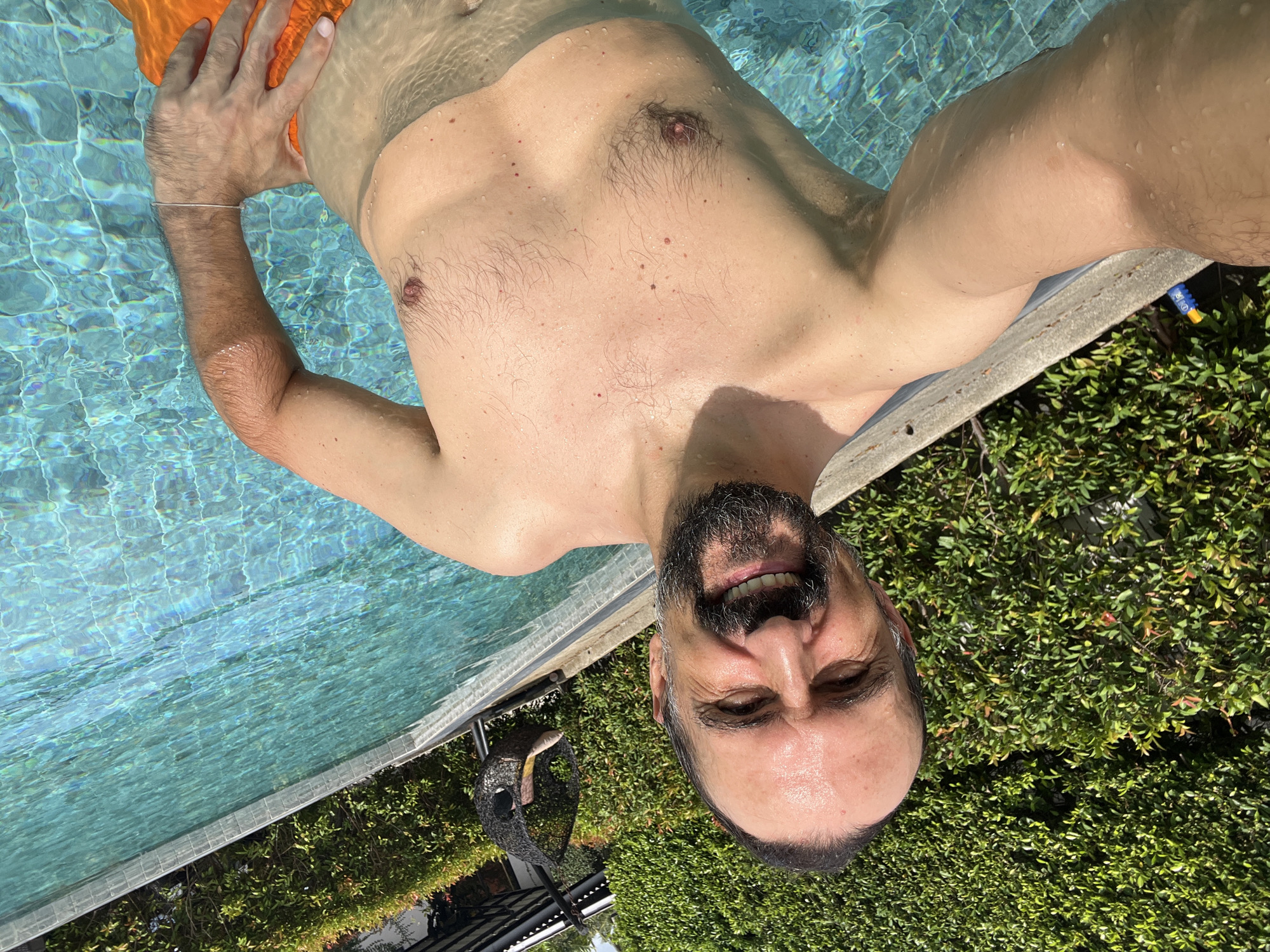 A man is partially submerged in a swimming pool, likely in บ้านบางรักษ์, Thailand. He is wearing orange swimming trunks and appears to be taking a selfie. The pool has light blue water and a tiled edge. Beyond the pool edge is green foliage, possibly hedges or shrubbery. A decorative object or sculpture can be seen near the edge of the pool and the foliage. The man is smiling and has a beard.