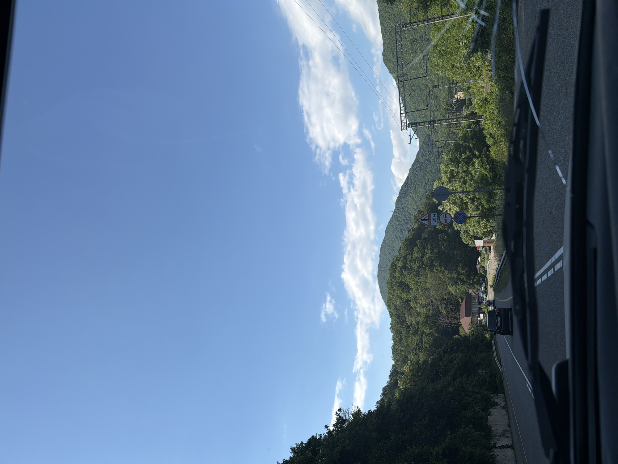A forward-facing, eye-level shot captures a scenic view from inside a vehicle on a clear day, overlooking a road winding through a lush, mountainous landscape in Tuapse, Russia. The sky is a vibrant blue with scattered white clouds, and the sun illuminates the rolling green hills covered in dense trees. A highway, marked with double white lines, stretches into the distance, with a dark-colored car visible ahead. Power lines and poles are prominent on the right side of the road, indicating infrastructure in the area. A few road signs are also visible on the left, including one with "120 M" and a warning symbol. A house with a brown roof is nestled among the trees in the middle distance. The foreground shows the windshield and wipers of the vehicle.