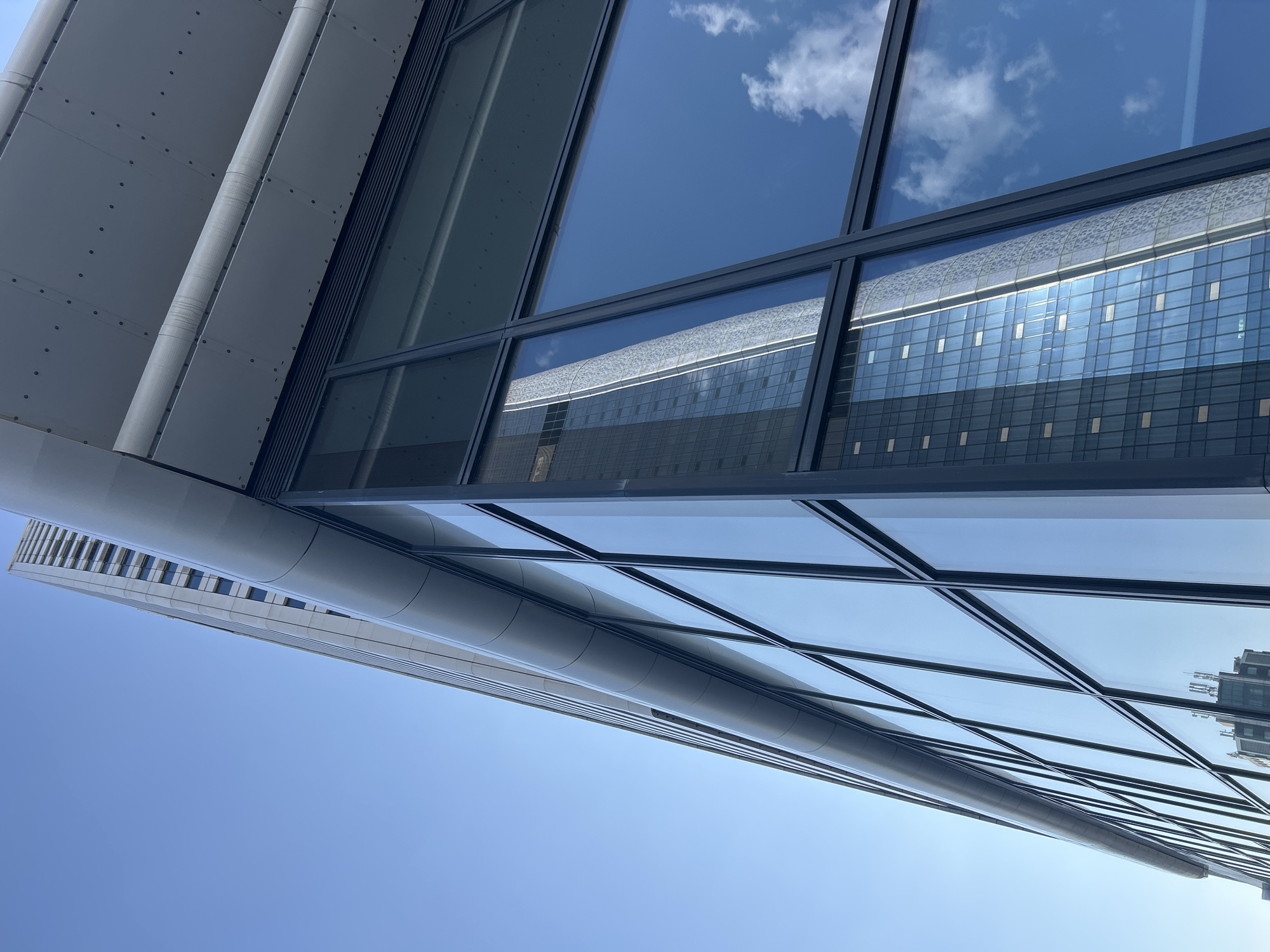 This is a low-angle, close-up, full shot of a modern skyscraper with a glass facade, reflecting a clear blue sky with sparse white clouds. The building's exterior features a sleek, contemporary design with clean lines and a metallic finish on its upper sections. The glass panels are segmented by dark frames, creating a grid-like pattern. In the reflection on the glass, another building of similar architectural style is visible, also appearing tall and modern. The overall impression is one of urban development and sophisticated architecture, set against a bright, sunny day. The specific location in Sofia, Bulgaria, is not visually determinable from the image alone.