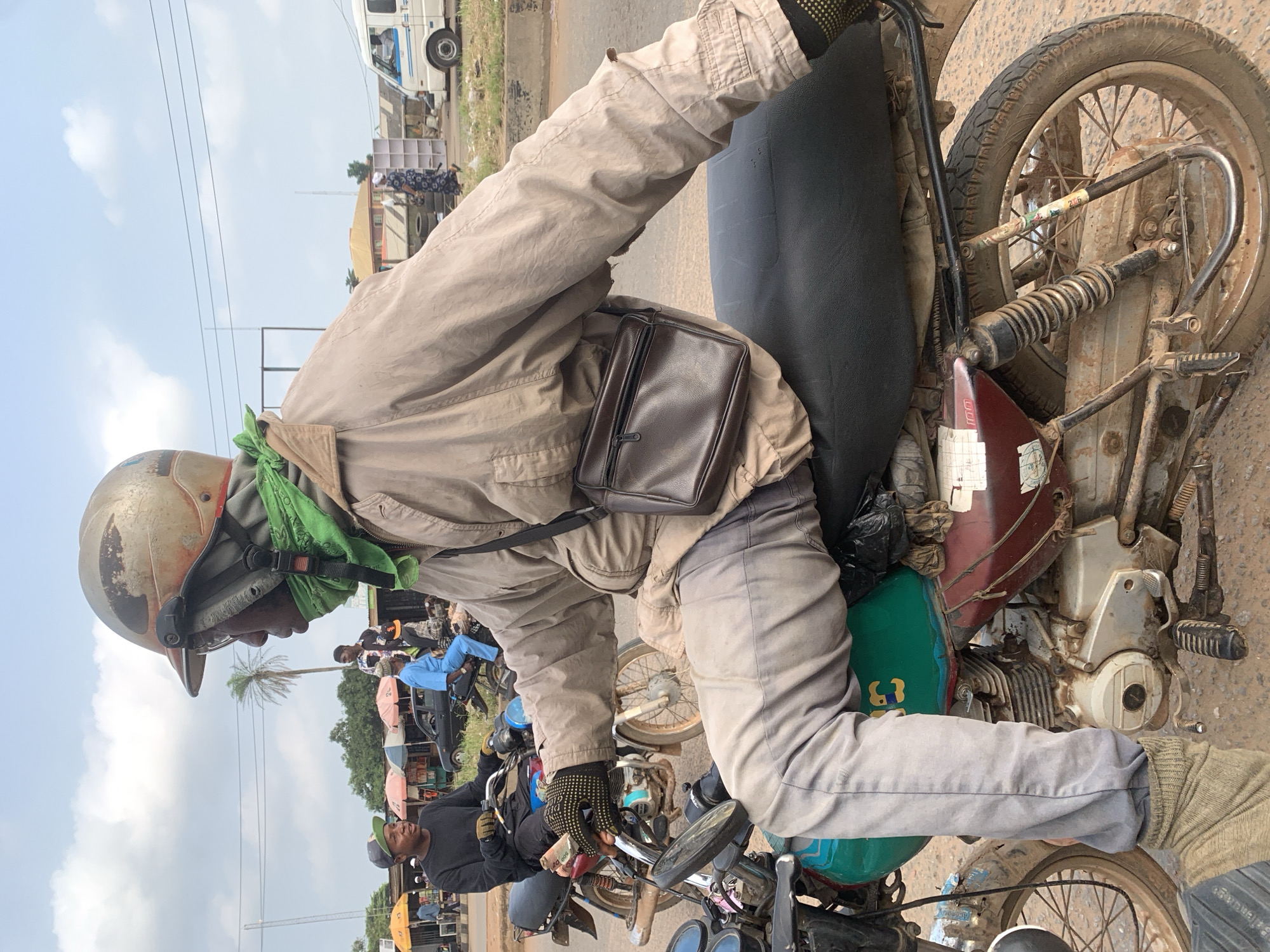 A medium shot captures a man in profile, seated on a motorcycle in Shagamu, Nigeria, on a dusty road. He wears a faded beige jacket, grey pants, and a worn helmet with rust and scratches. A green cloth is tied around his neck, and he has a small brown satchel slung across his chest. His gloved hands grip the motorcycle's handlebars, and he appears to be holding money. Another man is visible on a motorcycle behind him, and other people and market stalls are in the background. A white van drives by in the distance. The scene suggests a busy street market environment.
