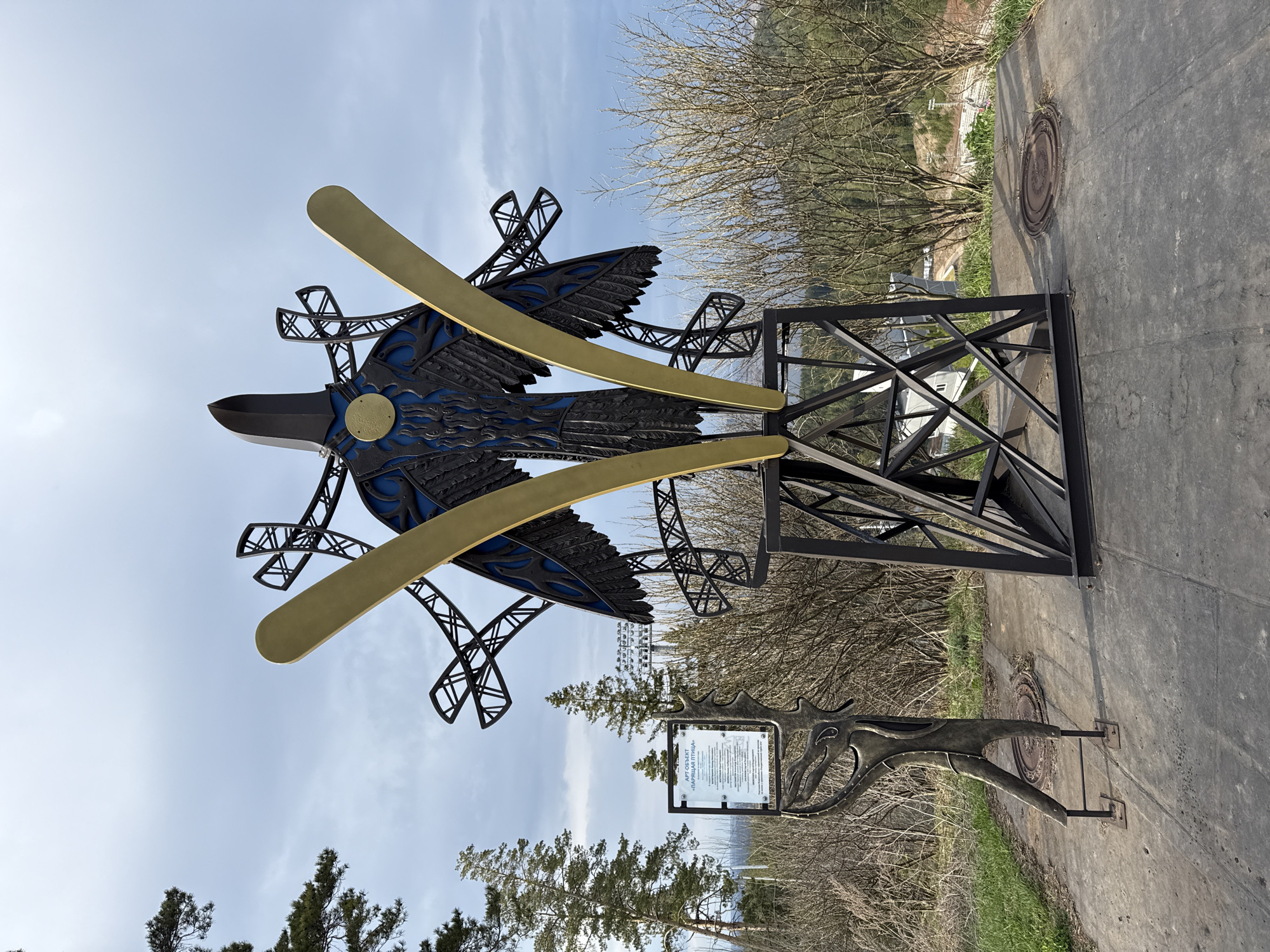 The image shows an outdoor art installation in Прикамский, Russia. The central element is a stylized sculpture of a bird with outstretched wings, constructed from black metal and accented with blue and gold. It appears to be mounted on a black metal frame. To the right of the sculpture, there's a manhole cover embedded in the pavement. Below the bird sculpture, a decorative metal sign with a plaque is present, partially obscured by greenery. The background consists of trees, bushes, and a partially visible urban or industrial landscape. The sky is overcast with visible cloud cover.