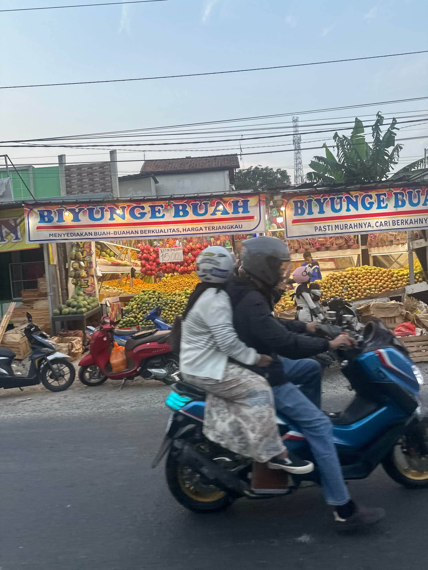 In Saruni, Indonesia, a street scene unfolds with a fruit stand as the focal point. The stand, adorned with a prominent banner proclaiming "BIYUNGE BUAH" and offering "MENYEDIAKAN BUAH-BUAHAN BERKUALITAS, HARGA TERJANGKAU" (Providing Quality Fruits, Affordable Prices) and "PASTI MURAHNYA, CARI BERKAHNYA" (Surely Cheap, Seek Blessings), displays an abundant array of fruits. Oranges are piled high, alongside green mangoes and pineapples. Behind the fruits, a person in a blue shirt can be seen standing.
In the foreground, two individuals are riding a blue motorcycle. The passenger, a woman wearing a white top and a patterned skirt, sits behind the male driver who is wearing a dark jacket and jeans. Both are wearing helmets, one grey and one white with a grey visor. Their motorcycle is in motion on a paved road, suggesting they are passing by the fruit stand. To the left of the motorcycle, a red scooter is parked, and further to the left, a black and white scooter is also parked. The environment appears to be an urban or semi-urban setting with buildings and power lines visible in the background. The lighting suggests it is either early morning or late afternoon.