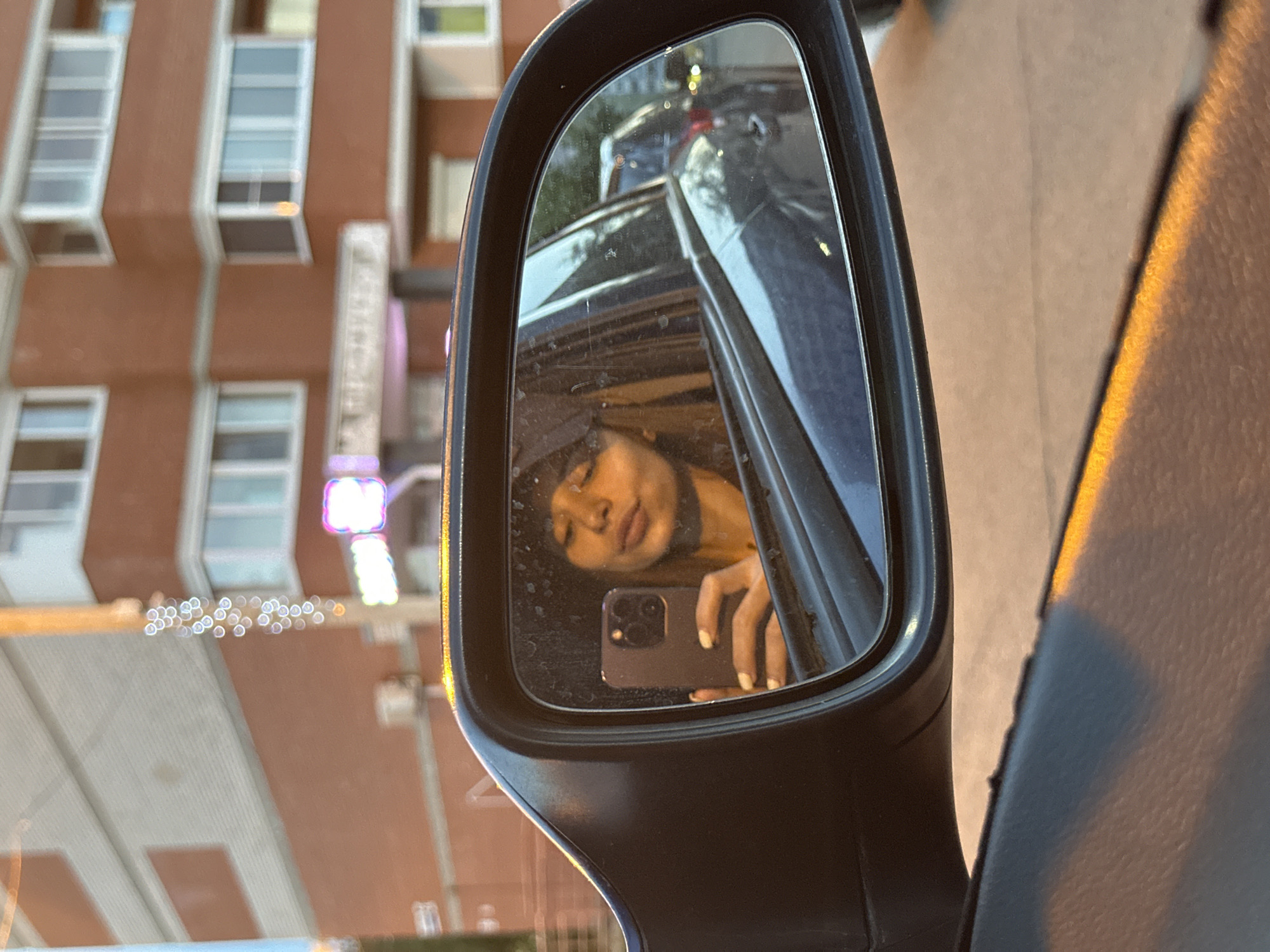 The image shows a side view mirror of a car, reflecting a person inside the vehicle taking a selfie with a smartphone. The person has dark hair, a fair complexion, and is wearing a hat. Outside the car window, a blurred view of a brick building with windows is visible. A sign with illuminated elements is also present on the side of the building. The location appears to be an urban setting with possible street lighting. The city, Izhevsk, Russia, cannot be determined from the image alone.