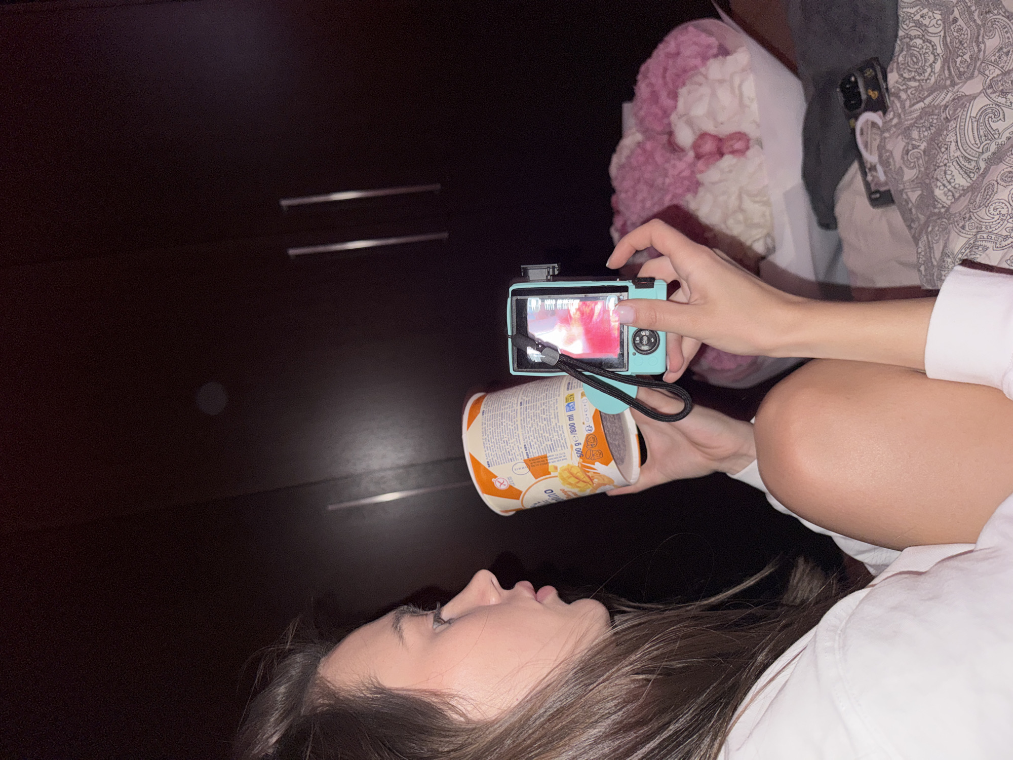 A photograph depicts a female subject lying on a dark surface, possibly a bed, next to a light pink stuffed animal, a duvet, and what appears to be a dresser. She is taking a photo with a small, blue digital camera of a cylindrical container with an orange lid. The container is labeled in a language that is difficult to discern, although the word “mango” is visible. The woman's hair is brown, and she is wearing a white shirt.
The photograph's location cannot be determined to be Warsaw, Poland, as there is no discernible evidence to support this claim in the image.