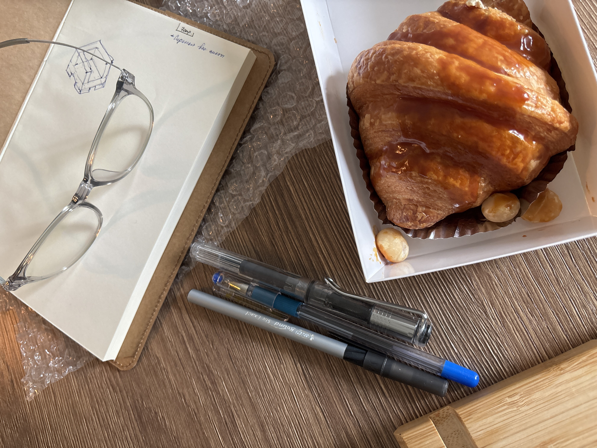 A still-life composition on a wooden surface. A notebook with a tan cover is open, displaying a drawing and handwritten text. A pair of clear-framed glasses rests on the notebook. A white box contains a glazed croissant and three macadamia nuts. Three pens are arranged near the box and notebook. The location is suggested by the prompt to be Choeng Thale, Thailand. Bubble wrap is partially visible in the background.