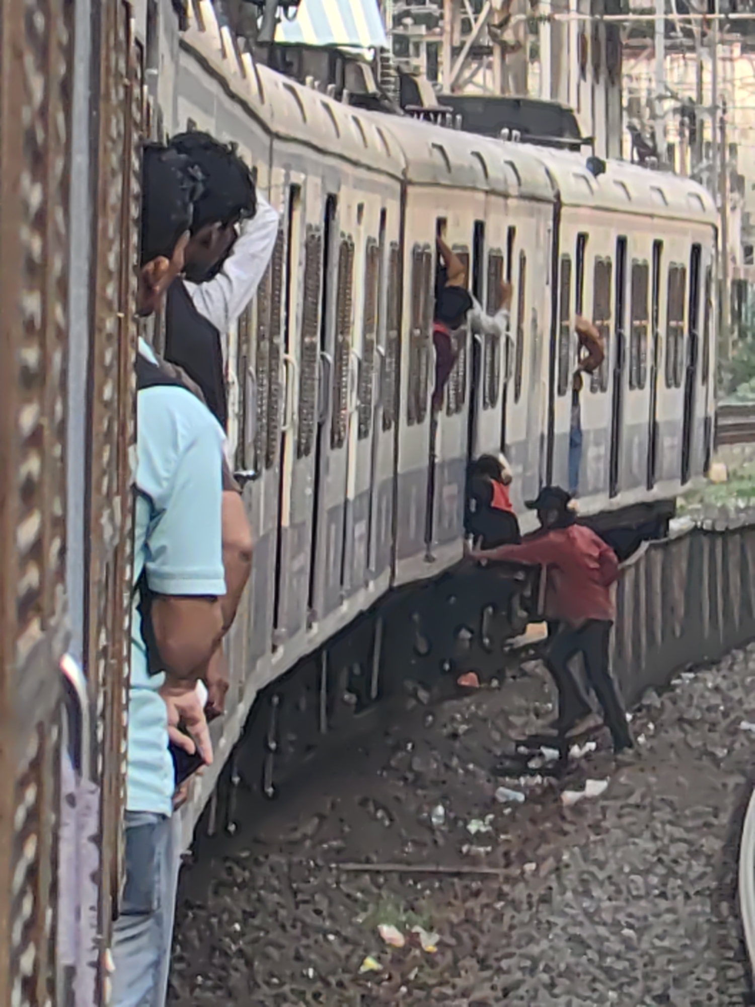 The image depicts a scene at a Mumbai suburban railway station, likely during peak hours. A densely packed train is visible, with individuals hanging out of the windows and doors. One person, wearing a light blue t-shirt and jeans, is leaning out of the train's door, holding a dark-colored mobile phone. Another person, in a red shirt and dark pants, is on the ground next to the train, reaching up towards the train. Several other individuals are partially visible in the train windows, some appearing to be young men. The train is stationary or moving slowly alongside a platform that is not fully visible. The ground beside the train is littered with debris, including plastic waste. The overall impression is one of overcrowding and potentially unsafe conditions on public transport in Mumbai.