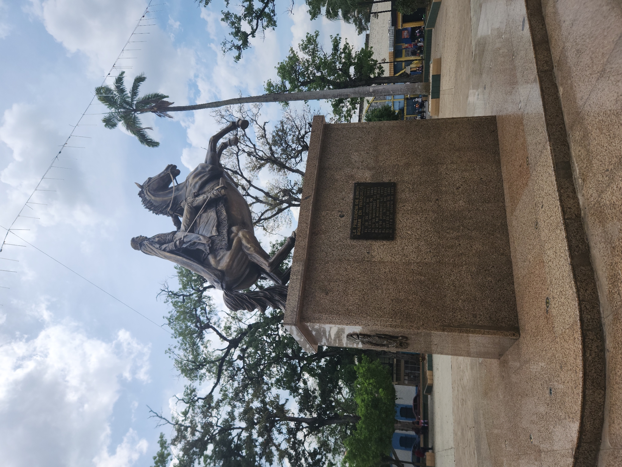 The image shows a bronze equestrian statue of Simón Bolívar mounted on a stone pedestal. The statue is in Tinaquillo, Venezuela, as indicated by the text on the plaque affixed to the pedestal: "LA PRESENCIA DE SIMON BOLIVAR EN TINAQUILLO". The plaque lists dates related to Bolívar's presence in Tinaquillo. A tall palm tree and other deciduous trees are visible in the background, with a partly cloudy sky above. Light strings are strung between the trees. The ground surrounding the statue and pedestal is paved. In the background, some buildings and people can be seen.