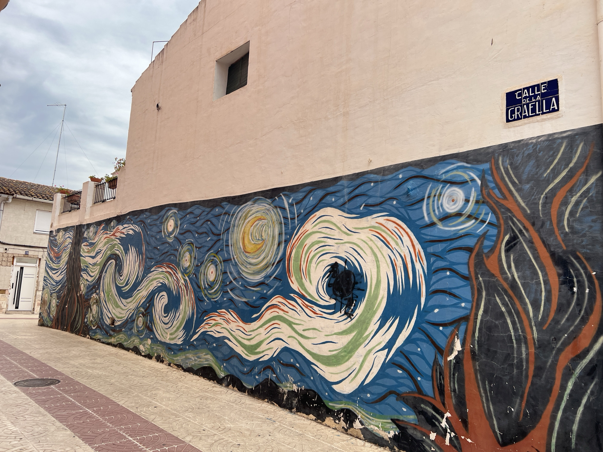 This image captures a street scene in Picassent, Spain, focusing on a long building wall adorned with a mural. The wall, painted in a light peach-beige, extends diagonally across the frame from lower left to upper right. A small, dark-shuttered rectangular window is set high on the wall's right side. Above the mural, on the upper right, a blue and white tiled street sign clearly indicates "CALLE DE LA GRAELLA." The lower portion of the wall features a detailed, colorful mural, approximately 1.5 meters tall, depicting an artistic interpretation of Vincent van Gogh's "The Starry Night." This mural showcases prominent swirling patterns in blue, white, green, and orange, against a dark blue background dotted with stylized stars. It includes a dark, gnarled tree-like structure with visible roots on the far left and a dark, flame-like or cloud-like form with orange outlines on the far right. The mural displays signs of deterioration, including peeling paint and cracking, particularly along its base where it meets the pavement. The street below is paved with light-colored tiles, segmented by two parallel strips of reddish-brown textured tiles extending into the distance. A circular metal manhole cover is visible on the pavement in the lower-left foreground. On the far left, a light-colored building with a white door, bearing a faint numeral "10" above it, is partially visible. Further back on the left, a building with a terracotta tiled roof and a balcony adorned with black metal railings and potted plants can be seen. The sky is uniformly overcast with light grey clouds. A tall, slender metal antenna with visible support wires rises into the sky on the upper left. No individuals or animals are present in the scene.
