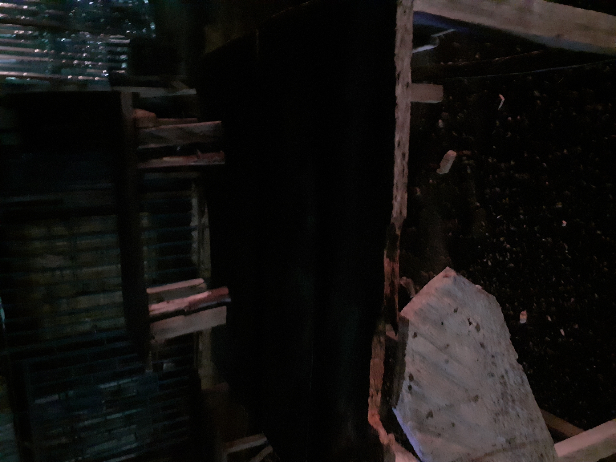 The image shows a dark, outdoor scene, possibly at night or in a poorly lit area. In the foreground, a large, irregular-shaped object, appearing to be concrete or stone, rests on the ground. It has a rough, textured surface with some debris scattered around it. Behind this object is a dark, flat surface, possibly a table or platform, supported by wooden legs. This platform extends into the middle ground of the image. In the background, there is a structure made of vertical wooden planks and a horizontal wooden beam, suggesting a fence or partition. The overall impression is one of a rustic or construction site environment. The image does not provide any specific clues to pinpoint its location within Jalingo, Nigeria, as the scene is generic.