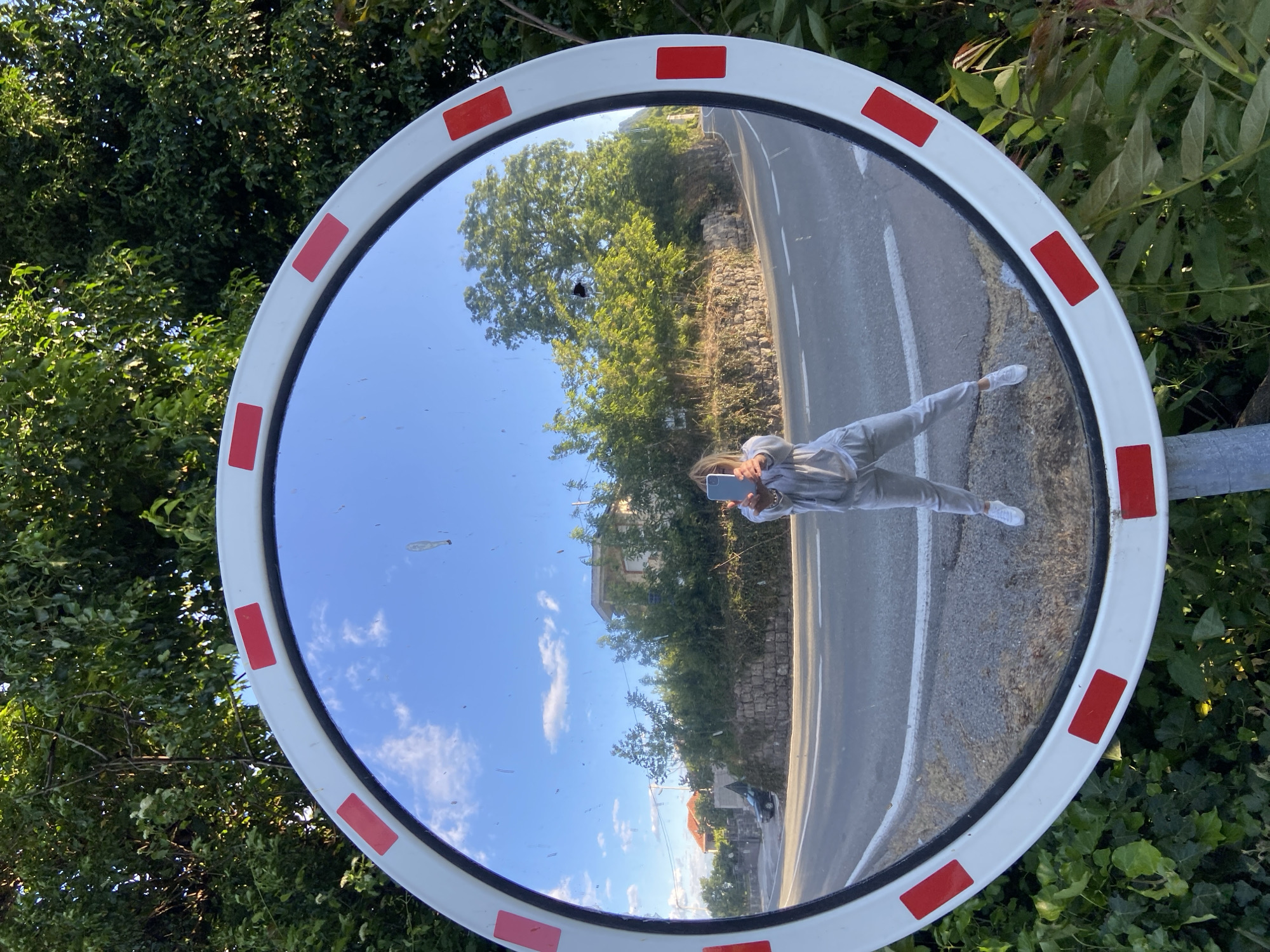 A large circular convex road mirror with a white frame and red rectangular reflective strips is the central subject of the image. Reflected in the mirror is a person, likely the photographer, taking a selfie with a smartphone. The person is dressed in light-colored casual attire, including a hoodie, pants, and white sneakers.
The reflection within the mirror shows a bright daytime scene. A paved road curves through the landscape, bordered by a rustic stone wall and abundant green trees and bushes. Beyond the immediate roadside foliage, a clear blue sky dotted with scattered white clouds is visible, along with distant buildings featuring white walls and terracotta roofs. The mirror itself has some smudges and dust marks on its surface.
The mirror is positioned amidst lush green vegetation, suggesting a rural or semi-rural roadside setting. The quality of light indicates fair weather conditions during the day. The architectural style of the distant houses and the general landscape are consistent with the location context of Sotonići, Montenegro. There is no visible text in the image.