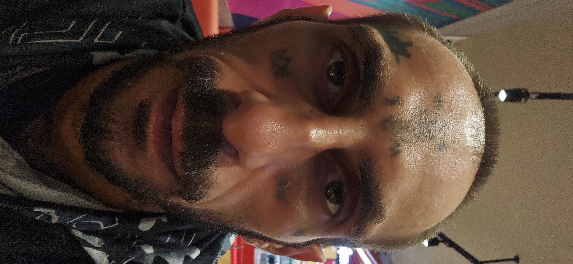 A close-up portrait of a man with facial tattoos, a beard, and short hair. The background includes elements of an interior space with possible lighting fixtures and red and pink accents, the image may have been captured in Bắc Nha Trang, Vietnam.
