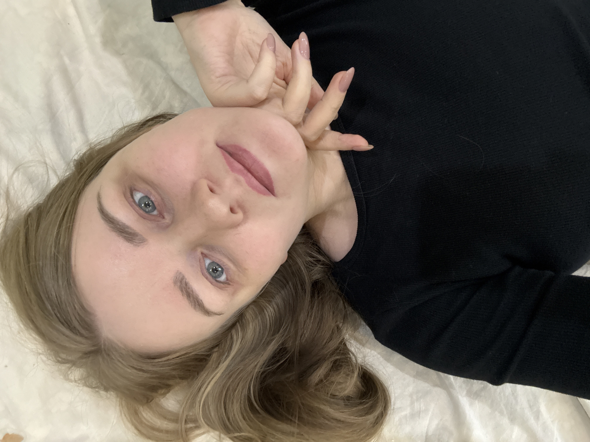 A young woman with fair skin, light brown hair, and blue eyes lies on a white surface, possibly a bed, wearing a black top. Her head is tilted, and her hand is near her face. Her fingernails are long and painted a light pink. The setting could be inside, but the provided image does not contain sufficient information to confirm its location in Chaikovsky, Russia.