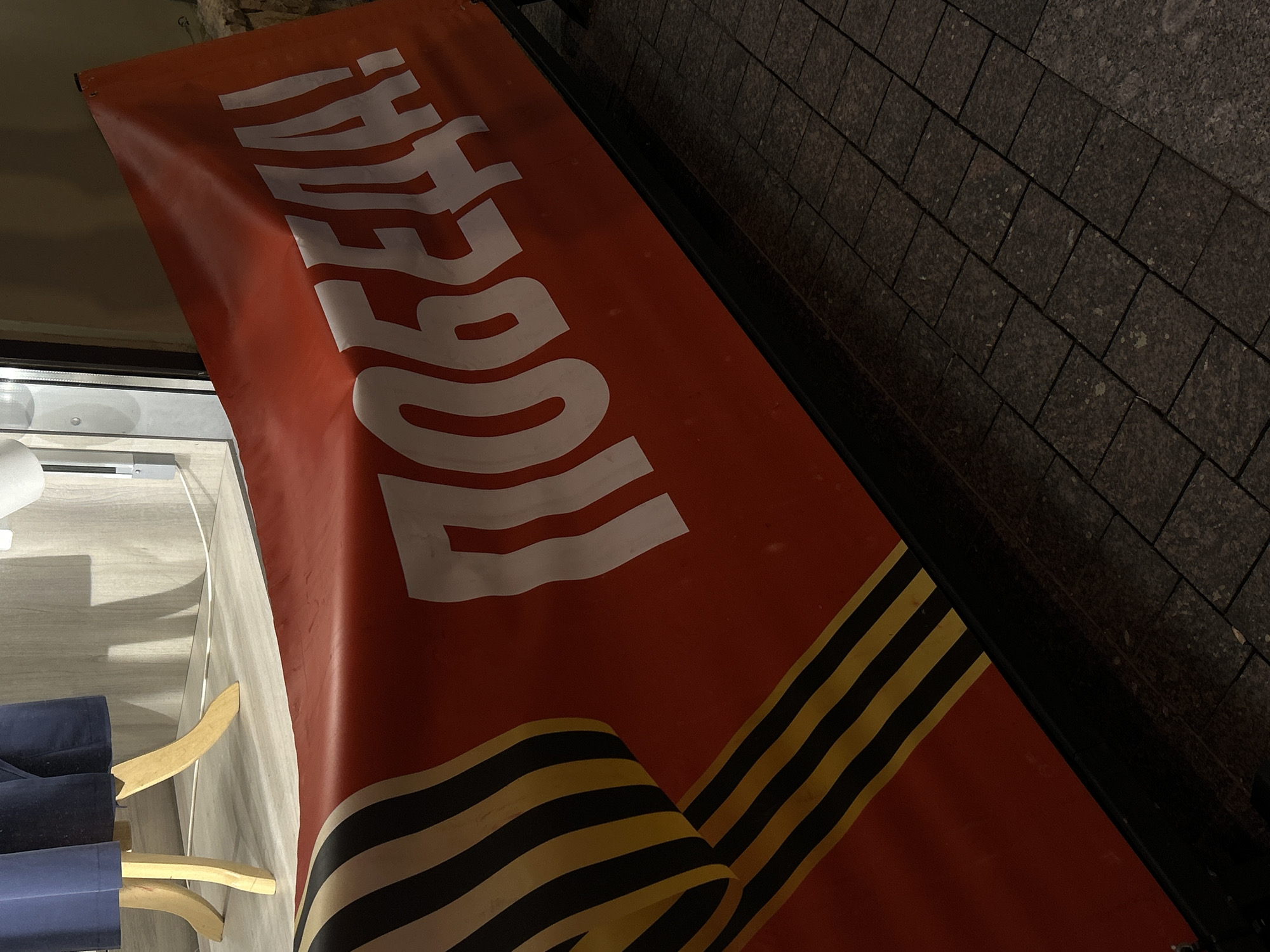 The image captures a diagonal view of a large red banner, likely displayed outdoors or in an open-air setting in Saint Petersburg, Russia. The banner features bold white Cyrillic text that reads "70 ЛЕТ!" (70 LET!), translating to "70 YEARS!". Below the text, a prominent graphic of a St. George ribbon is depicted, characterized by its black and orange stripes, suggesting a commemoration related to military victory, specifically the Victory Day celebrations in Russia, which would mark the 70th anniversary of the end of World War II (May 9, 2015).
The banner is situated above a partial view of what appears to be an indoor or semi-enclosed space, showing a dark blue upholstered chair with light wooden legs, a light-colored floor or surface, and a white object, possibly part of a table or another piece of furniture. To the right and upper part of the image, a dark, textured surface resembling a cobbled pavement or a tiled building facade is visible, separated from the banner by a dark beam or frame. The lighting is dim, suggesting it might be evening or night, or an interior with subdued artificial light. No people are visible in the frame. The overall scene conveys a sense of public commemoration within an urban environment.