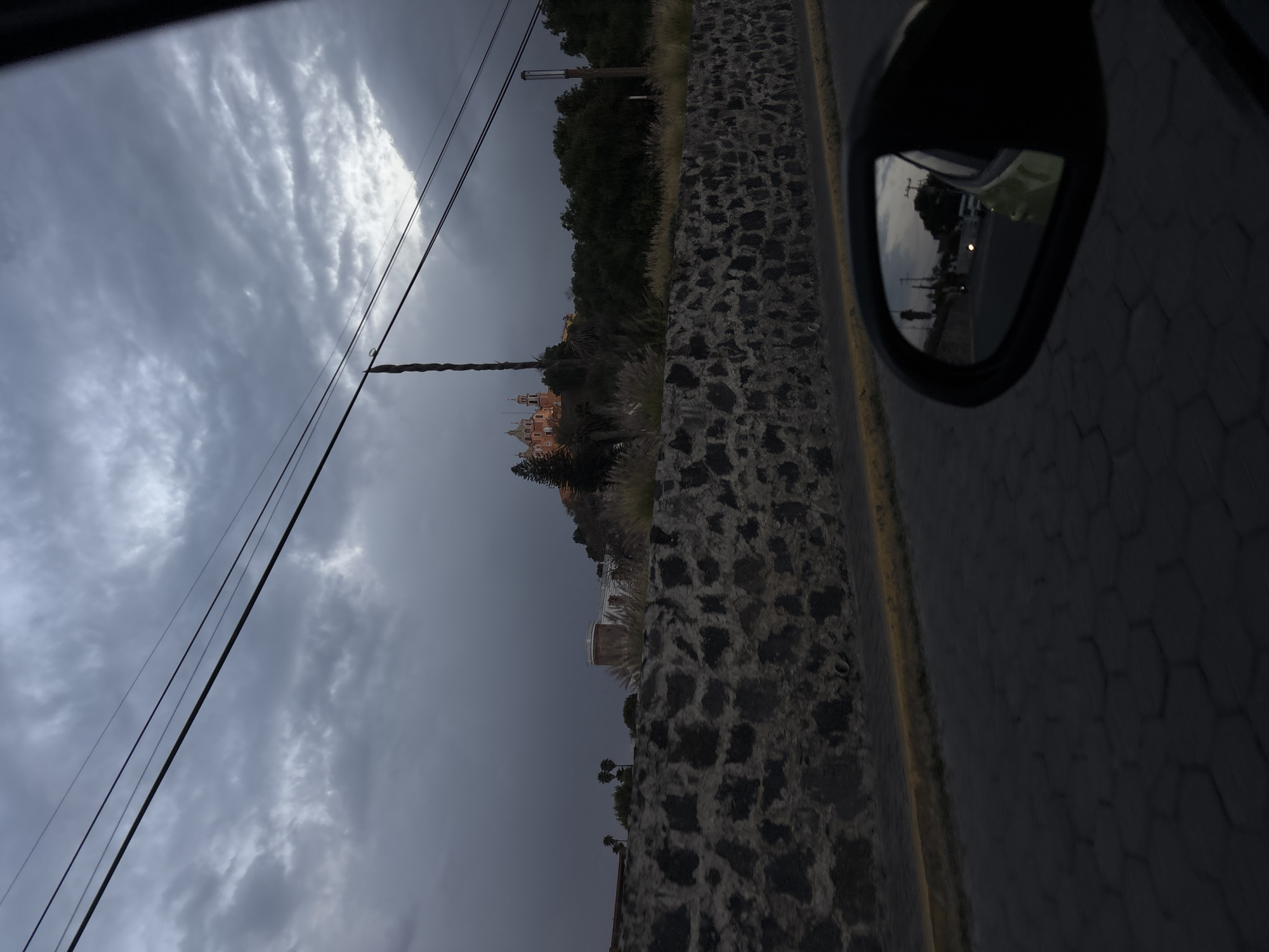 The image depicts an exterior scene in San Andrés Cholula, Mexico, at dusk or under heavily overcast conditions. The perspective is from inside a vehicle, looking towards the right side of a road.
In the lower foreground, a dark hexagonal-patterned paved road is visible, bordered by a yellow curb line. Above the curb, a dark, irregularly-shaped stone wall extends horizontally across the frame. To the lower right, the passenger-side exterior mirror of the originating vehicle is visible, reflecting a light-colored (lime green or yellow) vehicle, additional vehicles, and indistinct street elements traveling behind.
Above the stone wall, an elevated terrain is covered with dry, light-colored grasses and dark green foliage, including several palm trees. In the midground, the Templo de Nuestra Señora de los Remedios (Church of Our Lady of Remedies) is prominently situated on a large, tree-covered mound, identifiable as the Great Pyramid of Cholula. The church features a reddish-orange facade and white domes, and appears to be artificially illuminated, making it the brightest subject in the frame. To the right of the church, another dark, tree-covered hill rises, with a vertical modern street lamp pole positioned on its edge. Multiple dark utility poles and overhead power lines traverse the upper portion of the image. The sky is dominated by dense, dark gray clouds, indicating a heavily overcast environment, with a small area of lighter cloud visible in the upper right. No identifiable individuals or specific human actions are discernible.