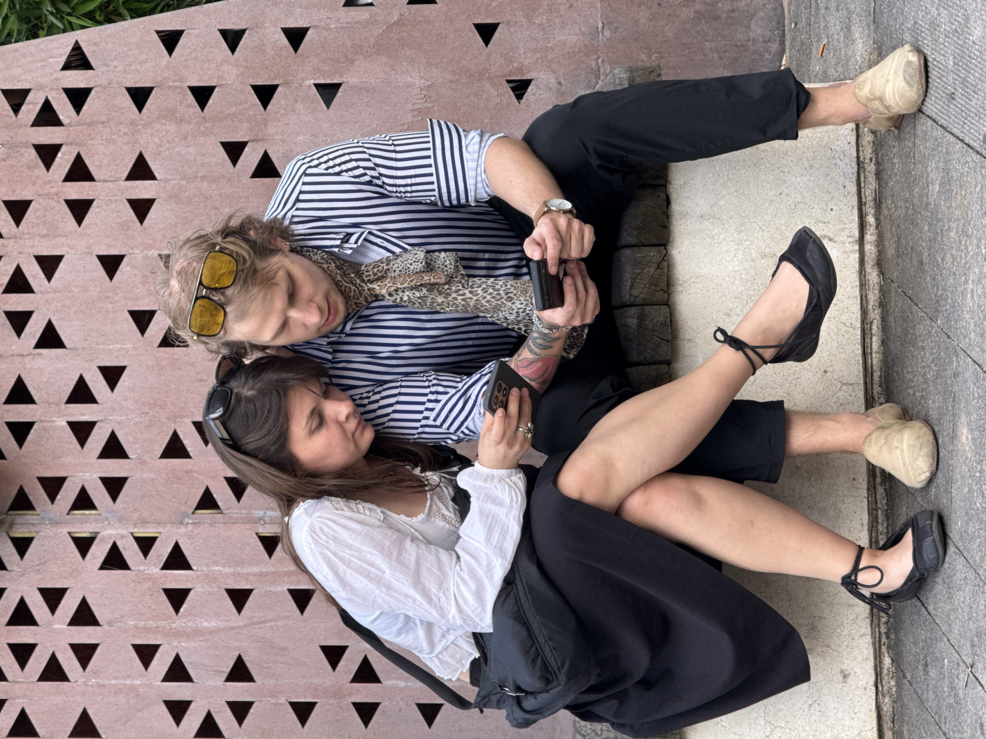 Two people are seated on a surface featuring a repeating triangular pattern. The individual on the left wears a horizontally striped shirt, a leopard-print scarf, and sunglasses resting on their head. They are holding a phone. The individual on the right is wearing a white shirt and black skirt and also holds a phone. They are seated closely together, with the person on the right resting their legs on the person on the left. The background surface has a pattern of triangles. The location appears to be in the Yuzhong District of China.
