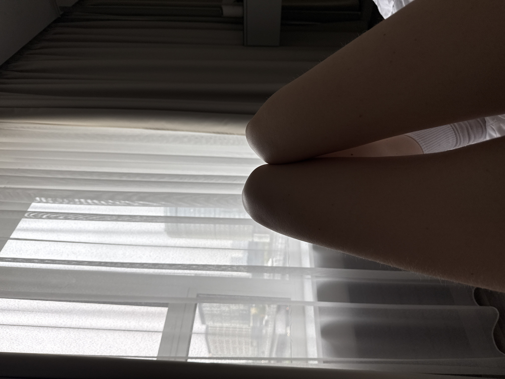 A close-up, interior shot depicts the crossed knees of an individual, positioned against a window. The window is covered with horizontal blinds, through which a cityscape is partially visible. The city is not identifiable. The lighting is subdued, casting shadows across the scene. The person is wearing a white garment that is visible between their knees. The overall composition is from a low angle, focusing on the knees and the partially obscured view outside the window.