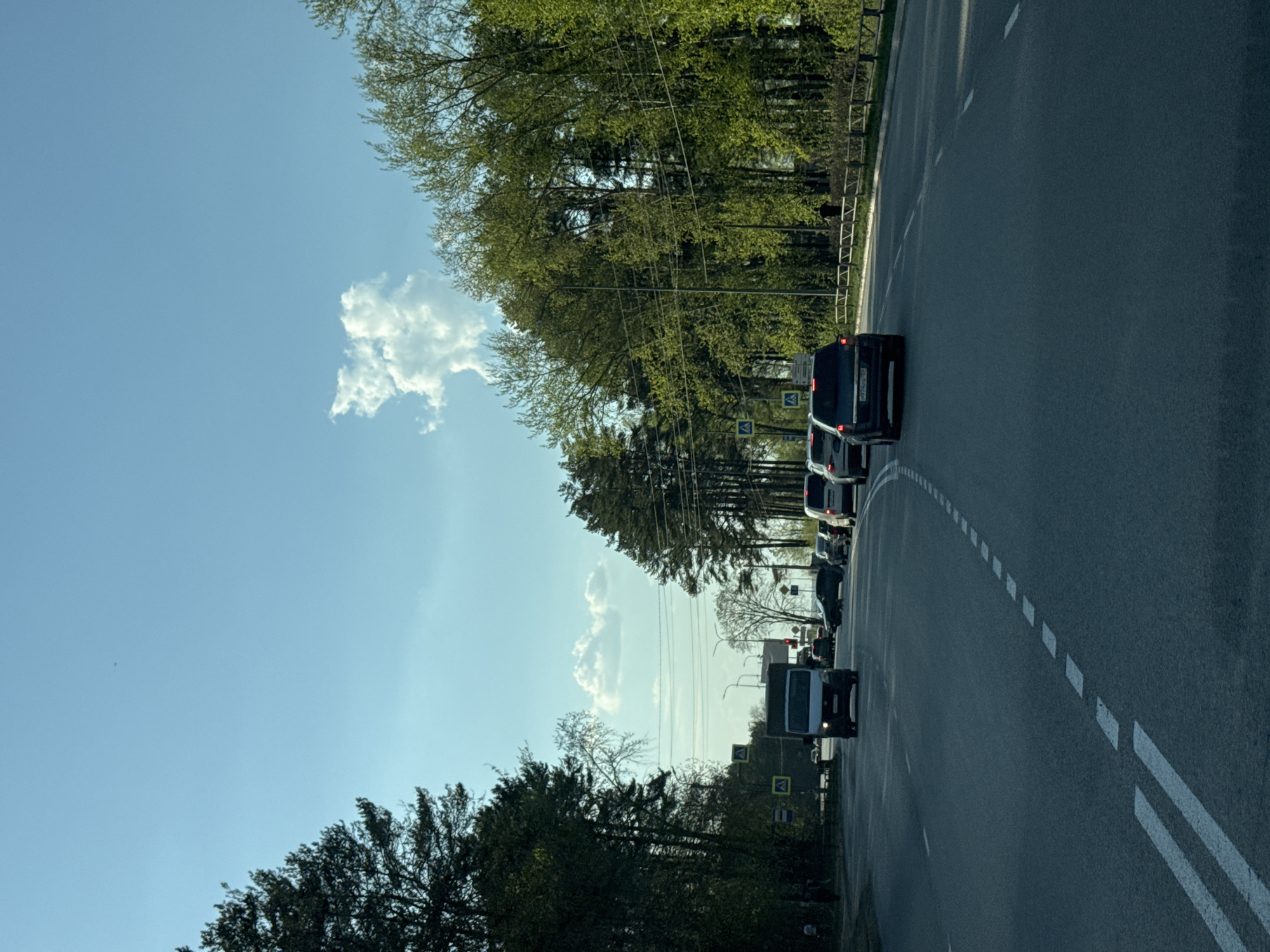 The image depicts a view from a vehicle on a road in Chaikovsky, Russia, heading toward traffic. The sky is clear blue with some clouds. Trees line the side of the road. Utility poles and wires are visible. The traffic includes a dark-colored van. Road markings consist of dotted and solid white lines. The time of day appears to be daytime.