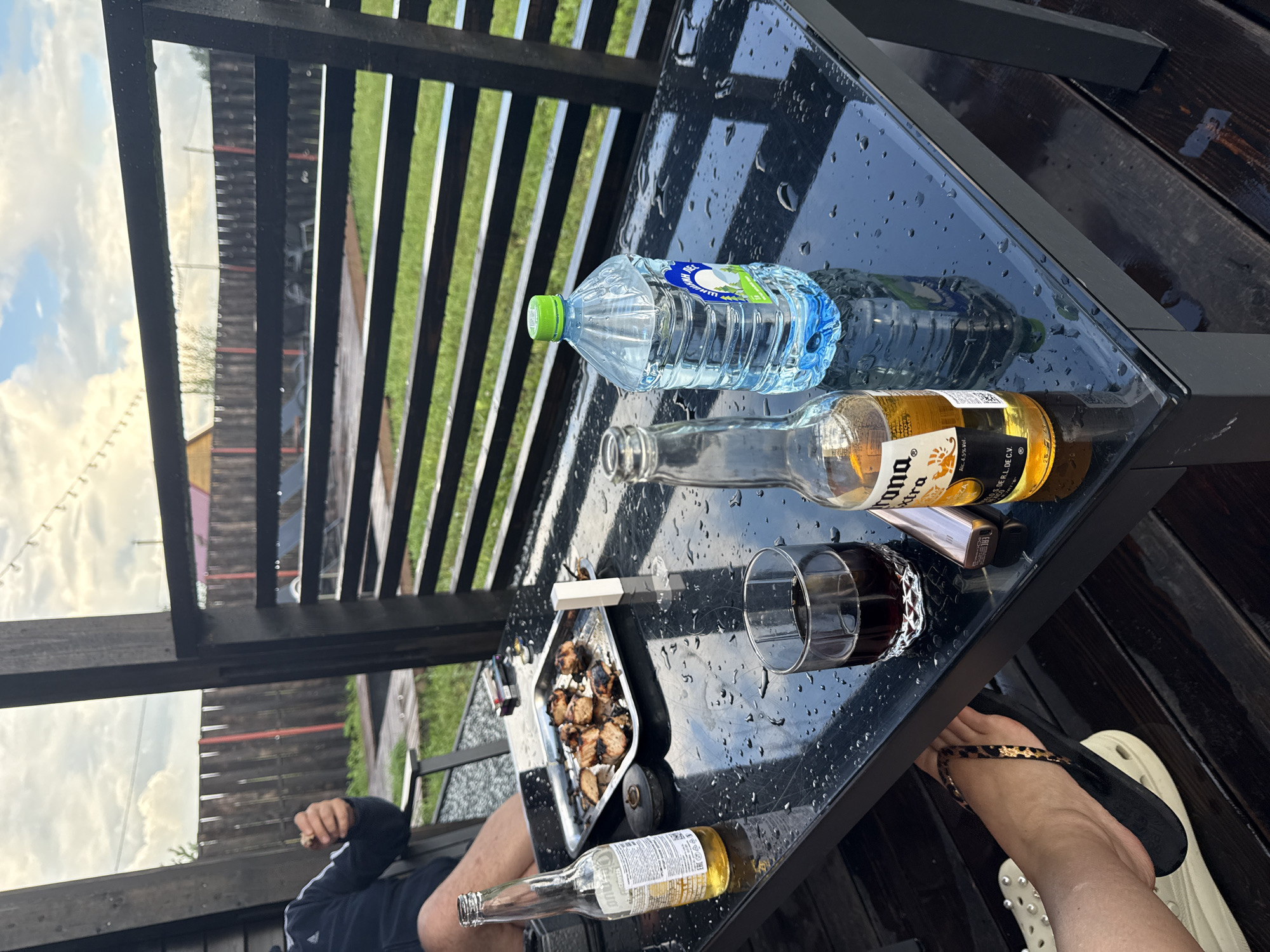 A person is sitting in a chair outdoors at a black table. The table has a bottle of Corona beer, a glass of dark liquid, and a bottle of water on it, along with a tray of grilled meat skewers and a lighter. The table and the wooden deck are wet, indicating recent rain. In the background, a wooden fence separates the area from a grassy yard. The sky is cloudy.