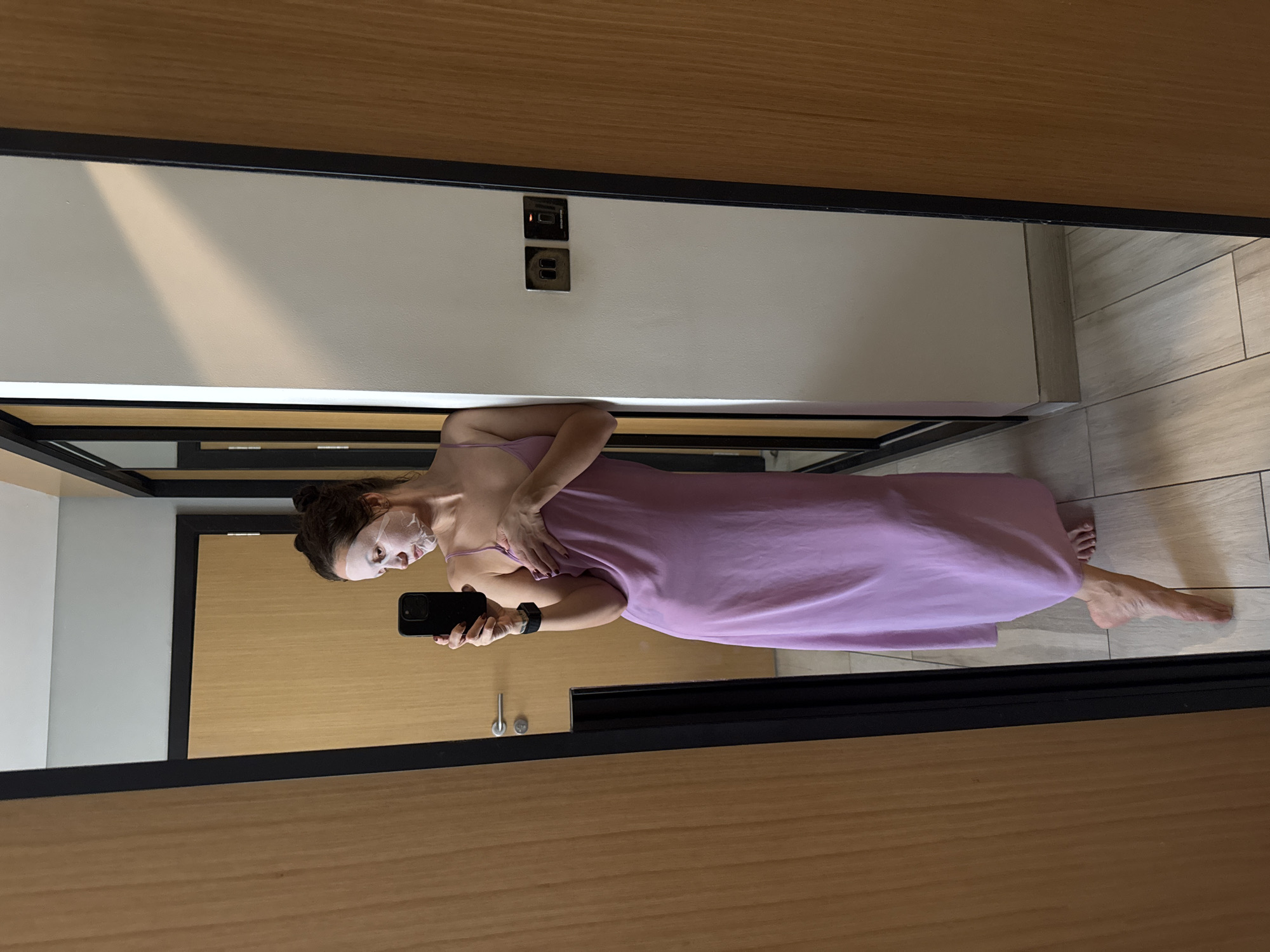 A woman is standing in front of a mirror, holding a smartphone in her left hand and taking a selfie. She is wearing a light purple slip dress and a white facial mask. Her hair is pulled back in a bun. The background shows a modern interior with light-colored walls, wooden doors, and tiled floors. There is a pair of electrical sockets visible on the wall above her. The location is inside a hotel or apartment, possibly in Dubai, United Arab Emirates given the style of the interior.