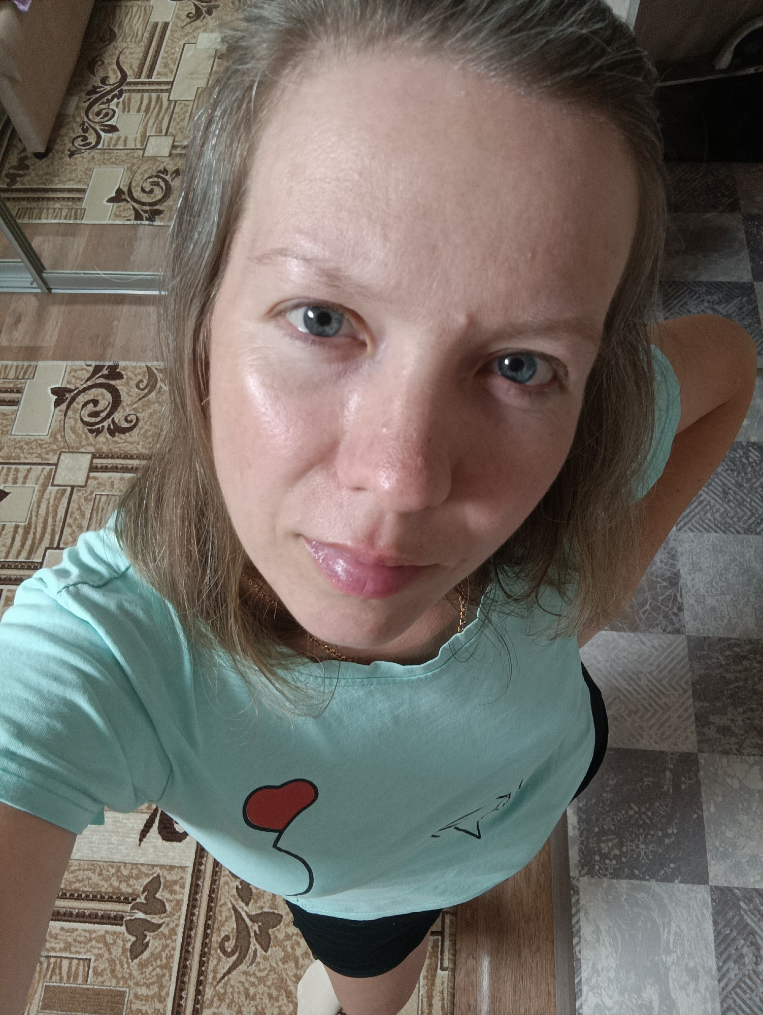 A middle-aged woman with fair skin, blue eyes, and light brown/graying hair is taking a selfie from a high-angle perspective indoors. She is wearing a light blue t-shirt with a red heart-shaped balloon design on the front. A gold chain necklace is visible around her neck. She appears to be standing on a floor with a patterned covering; one section has brown tones with a floral-like design, and another section has gray and white squares. Part of a metal-framed object is visible in the top left. The photo may have been taken in Chaikovsky, Russia.