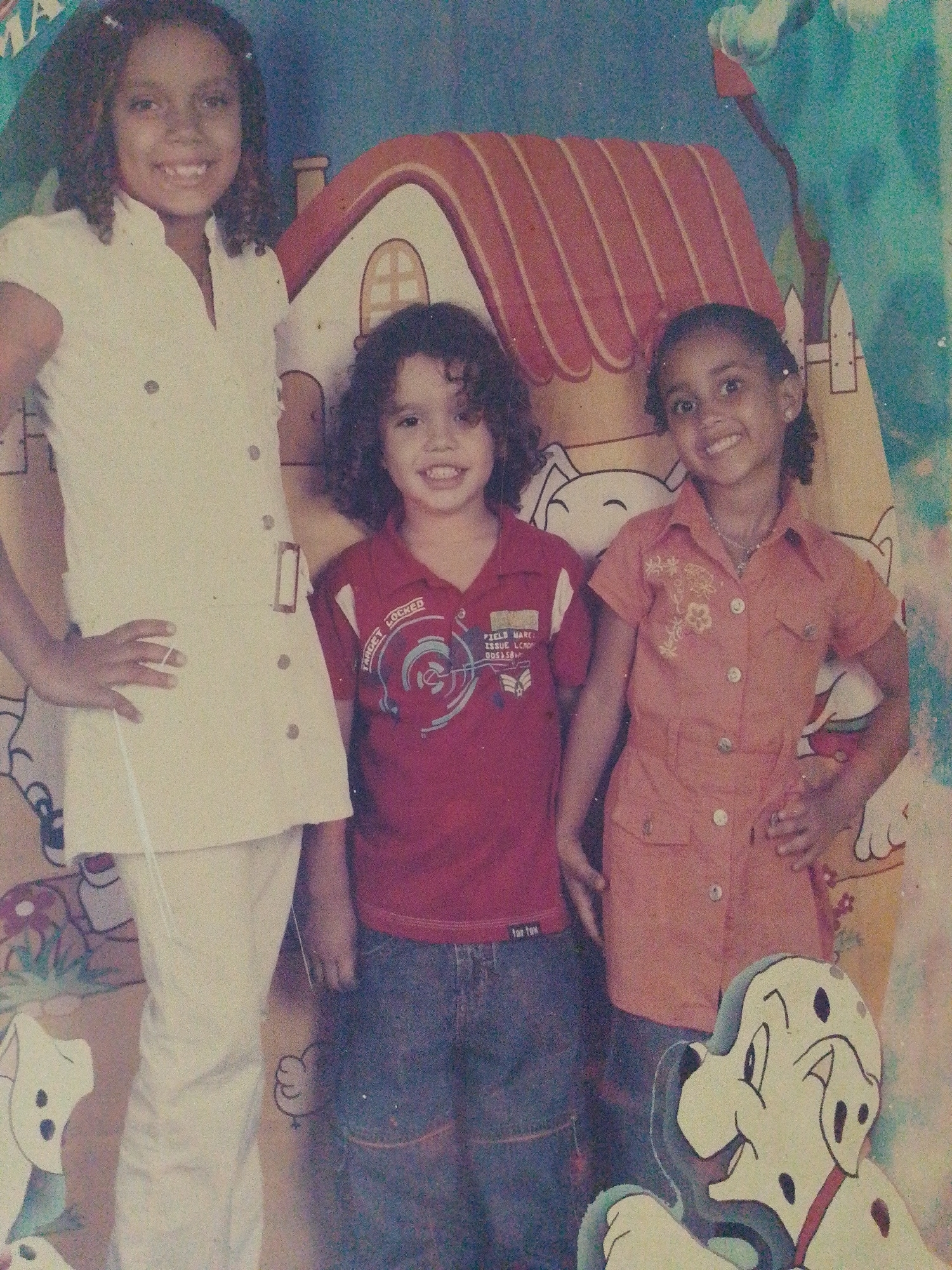 Three children are standing in front of a painted backdrop, possibly at a celebratory event in Tinaquillo, Venezuela. The child on the left is a taller girl wearing a white buttoned shirt and pants. The child in the middle is a boy with curly hair wearing a red shirt and jeans. The child on the right is a girl with braids wearing an orange buttoned dress. The backdrop features a cartoon house and characters, including a dalmatian.