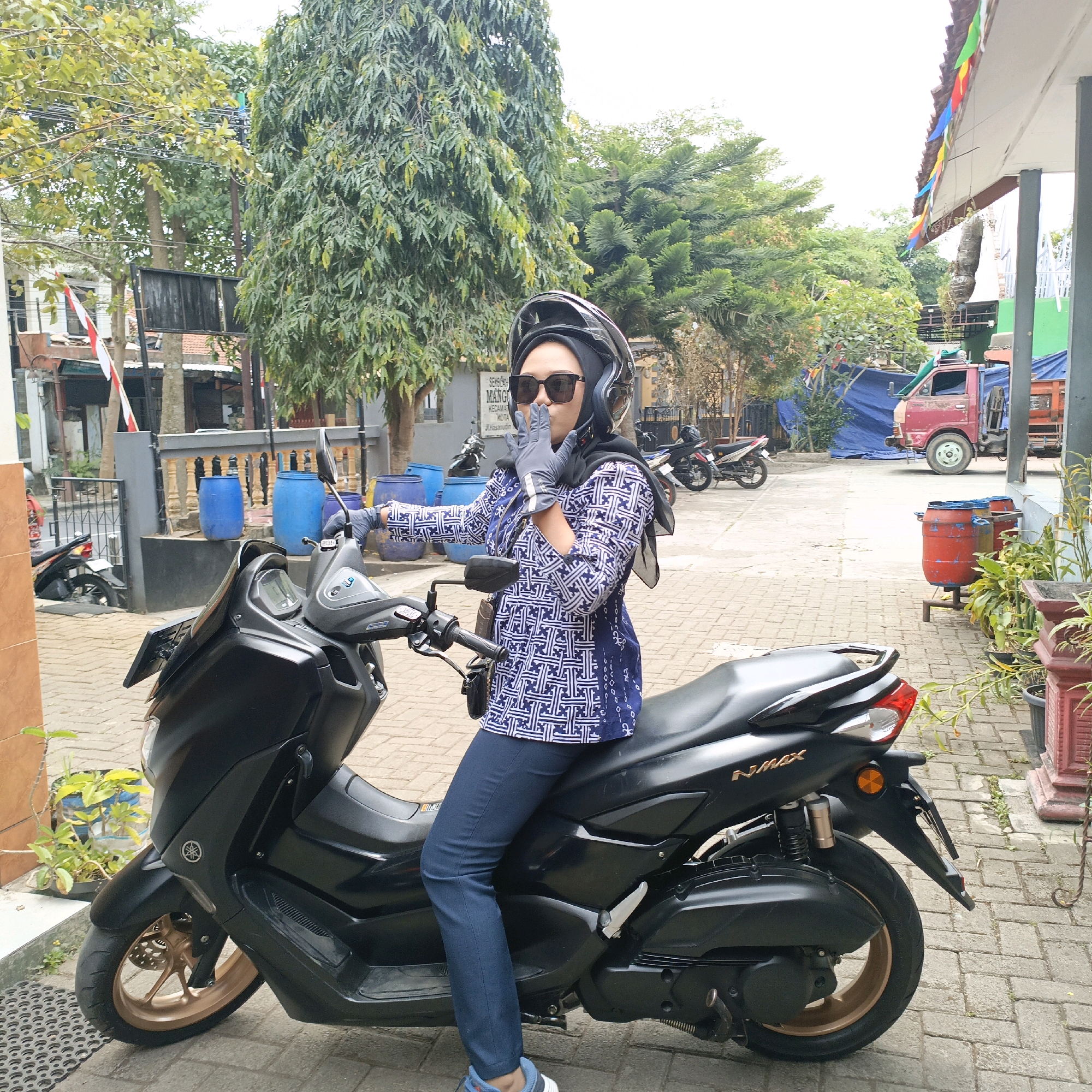 The image shows a woman sitting on a black Yamaha NMAX scooter. She is wearing a black hijab, a blue and white patterned long-sleeved shirt, dark blue jeans, a black helmet, and grey gloves. She is wearing sunglasses and has her hands up near her face, with her index fingers extended. The scooter is parked on a paved area that appears to be a courtyard or driveway. In the background, there are trees, a building with a red roof, several large blue barrels, a red truck, and other parked motorcycles. The location is identified as Salatiga, Indonesia, based on the sign visible in the background which reads "SEMANGGI KECAMATAN KOTA JL. HASANUDIN". The overall scene suggests an outdoor environment in a moderately urban or suburban area.